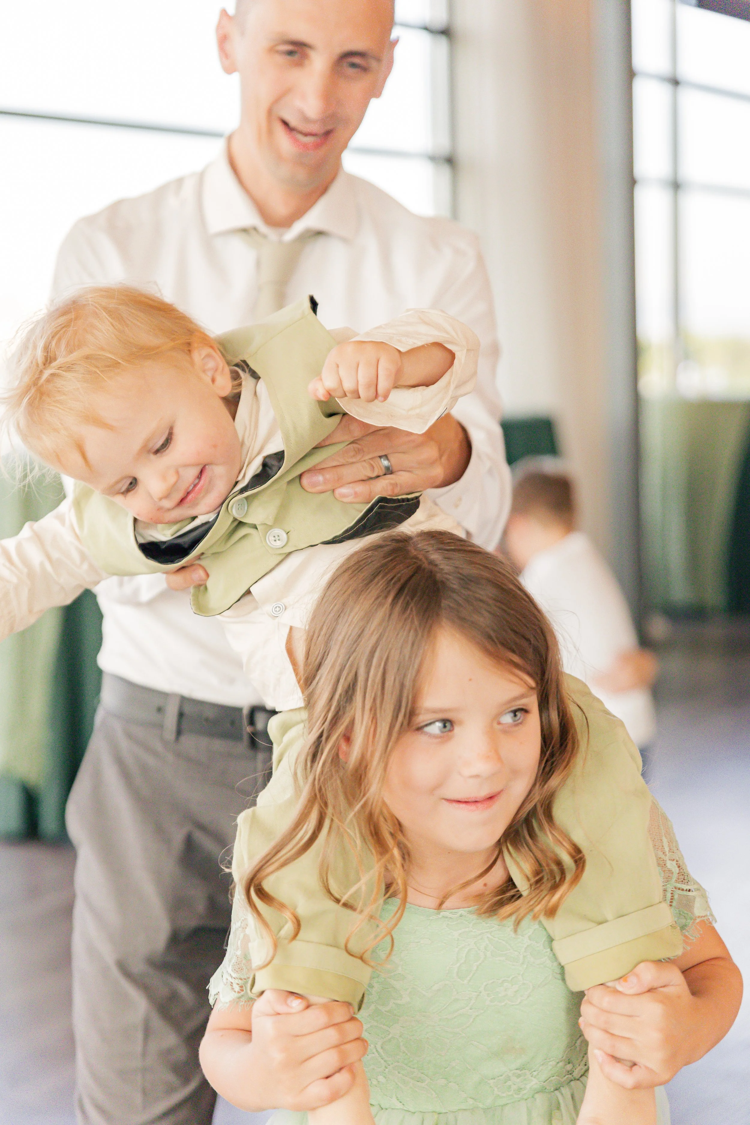 A man in a white shirt and beige tie holding and playfully lifting a young boy with red hair, while a young girl with light brown hair and a green dress stands in front, supporting the boy's weight on her shoulders, in a bright indoor setting.