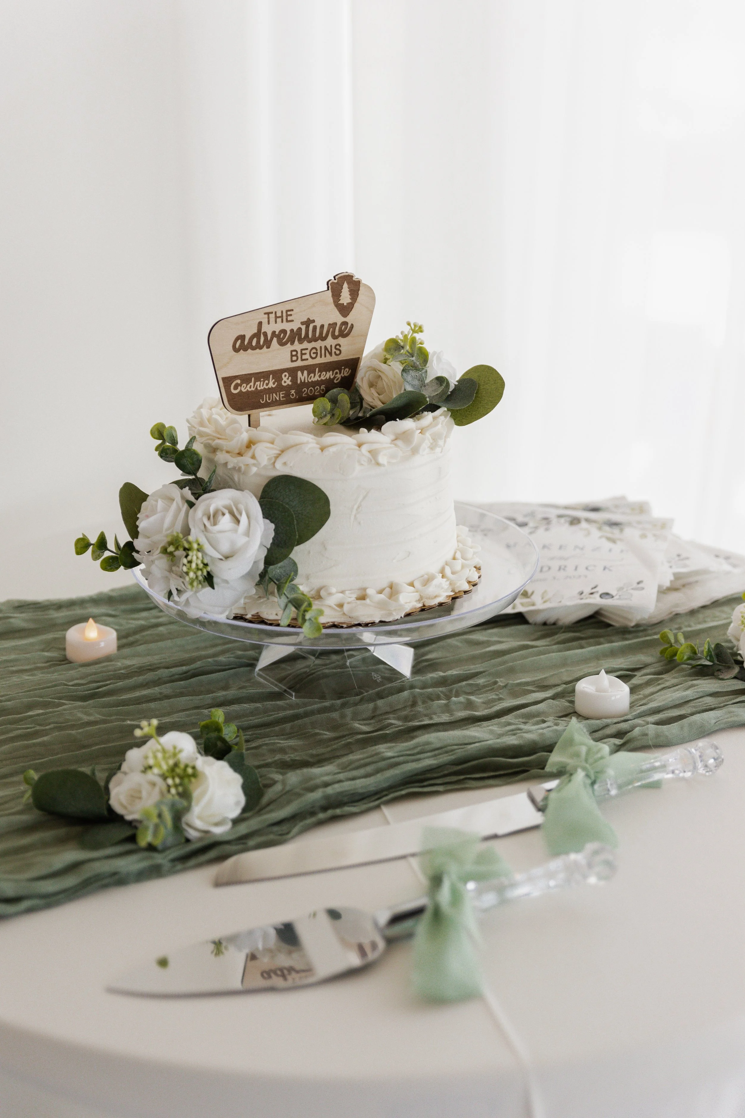 A white wedding cake decorated with greenery and white flowers, featuring a sign that says "The adventure begins Cedrick & Makensie June 3, 2025," placed on a glass cake stand. The cake is on a table with a green fabric runner, surrounded by small ca