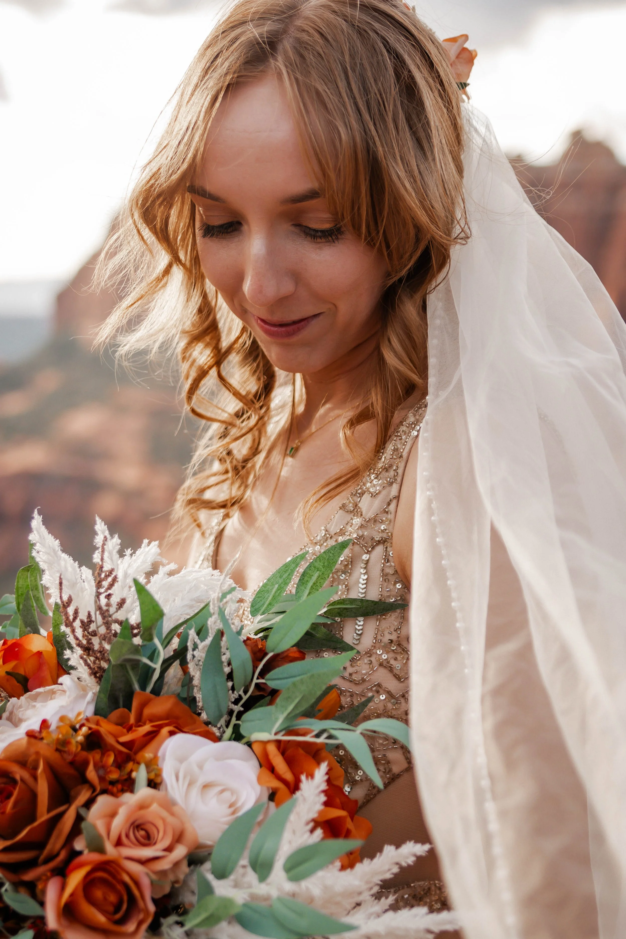 A woman with long, wavy, reddish-brown hair, wearing a beaded dress and veil, holding a bouquet of orange, white, and peach roses with greenery, outdoors with rocks and trees in the background.