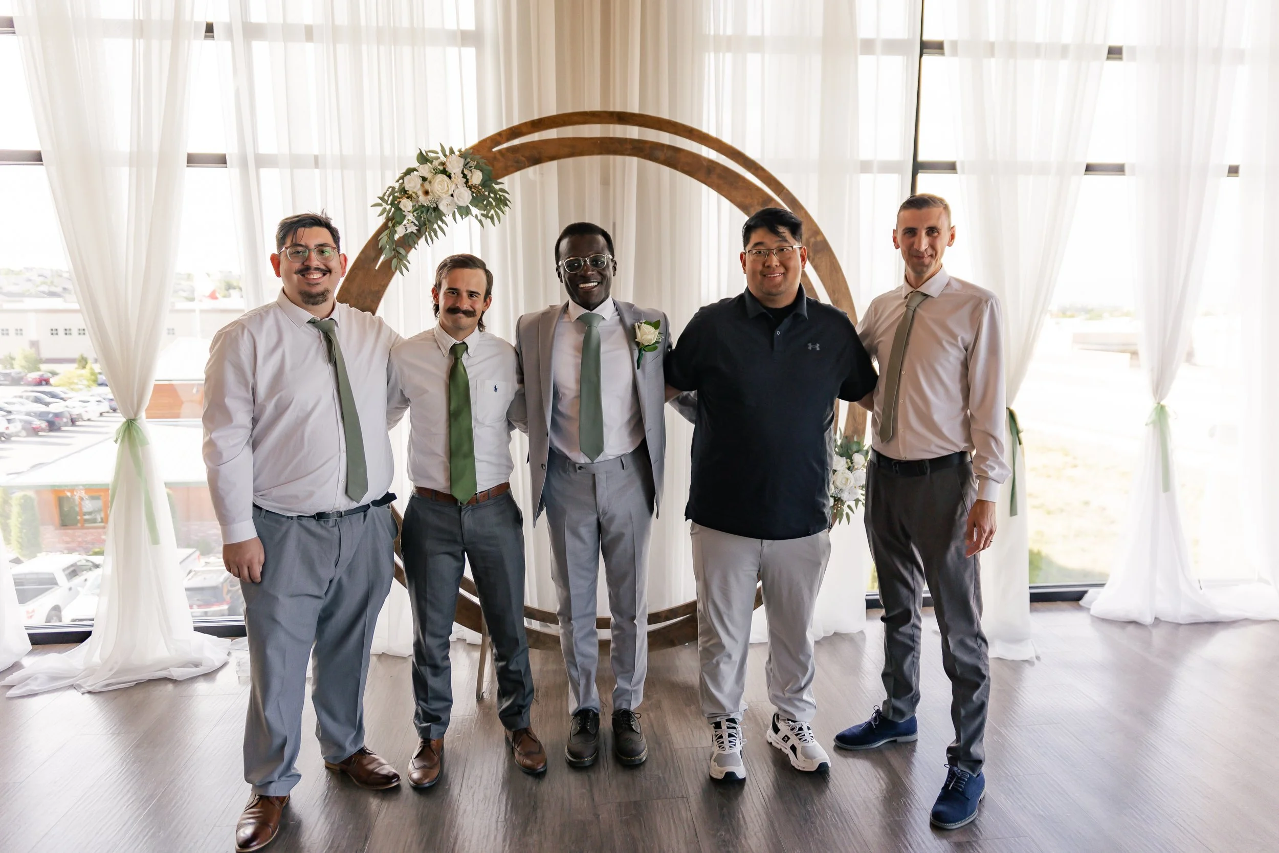 Group of six men standing together at a wedding or formal event, in front of a circular floral backdrop with white curtains and windows in the background.