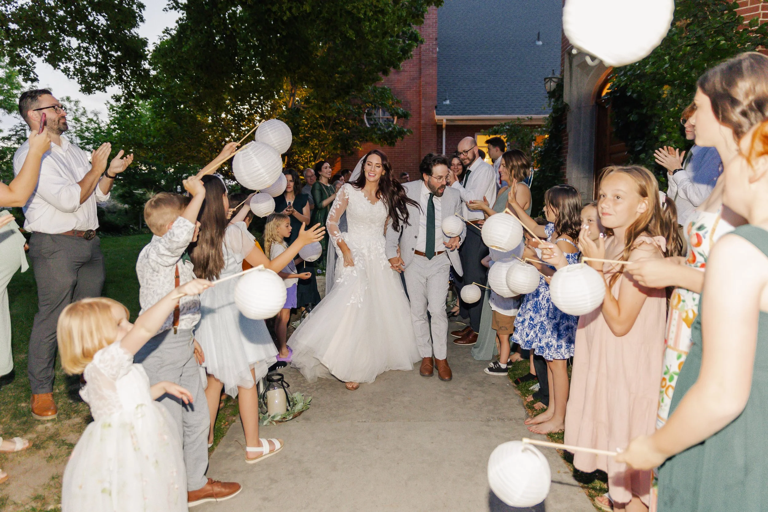 A wedding celebration outdoors with a bride and groom walking through a line of children and adults holding paper lanterns and clapping on either side.