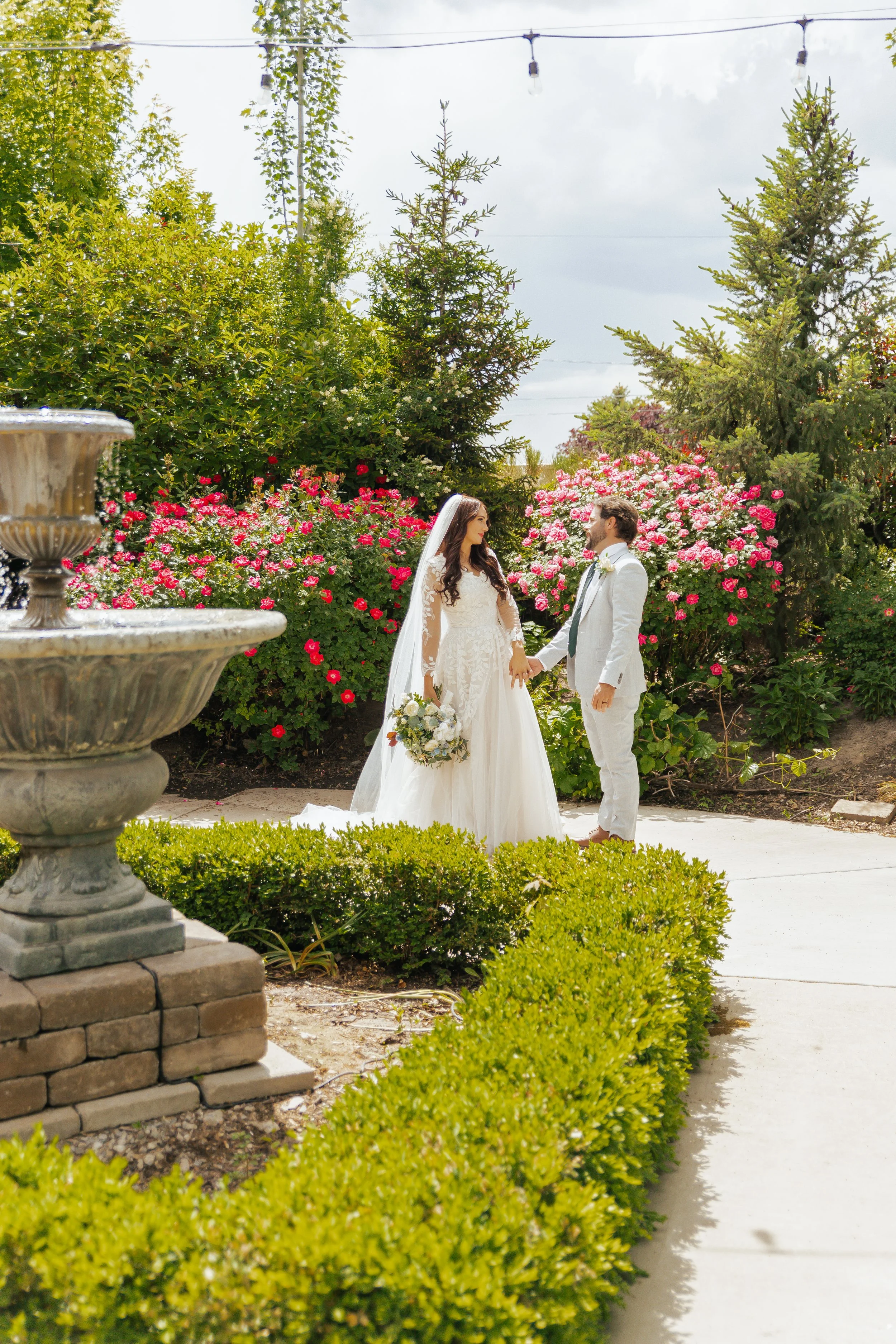 Bride and groom holding hands in a garden with pink flowers, green shrubs, and trees, during a wedding ceremony.