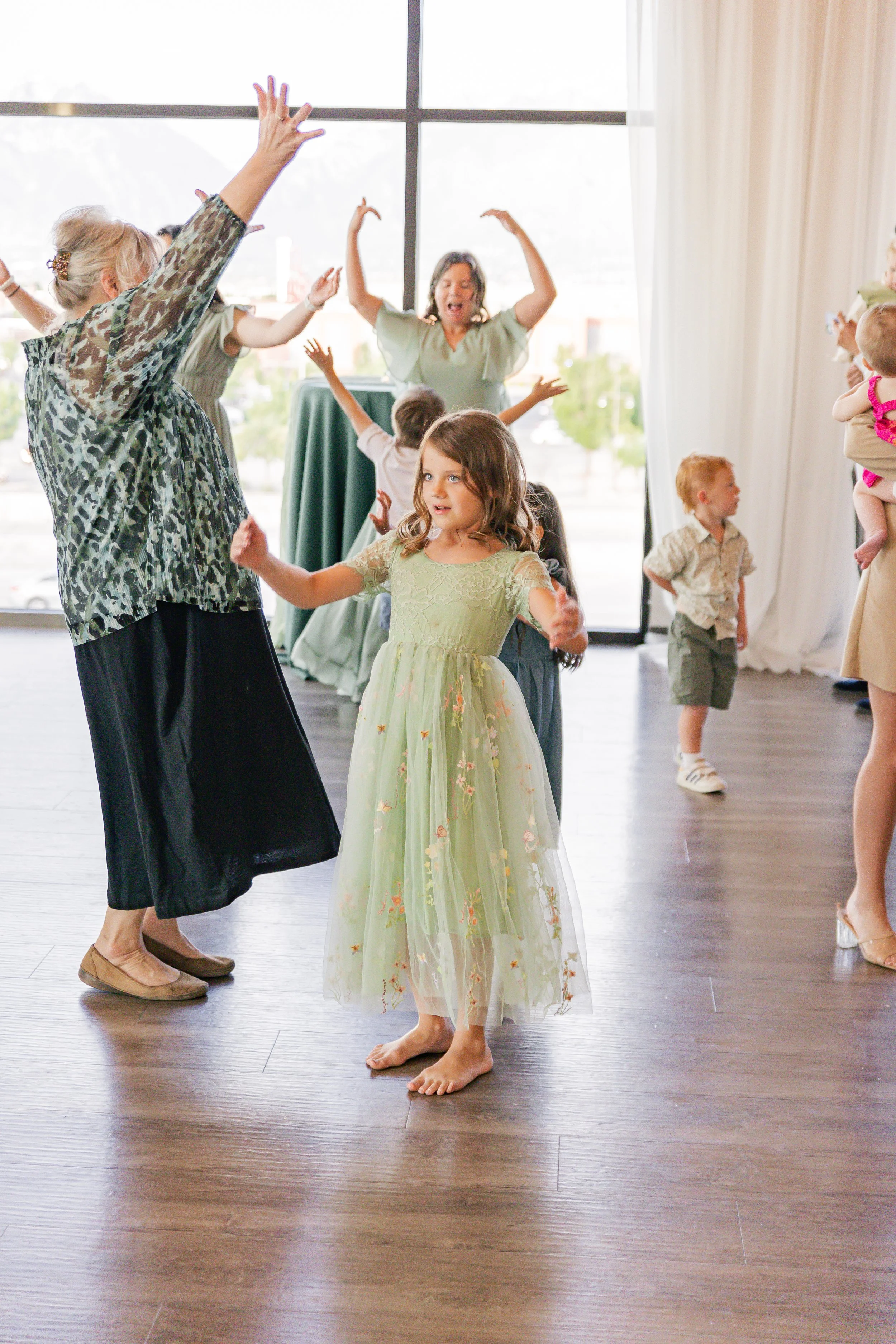 Children and adults dancing and playing together indoors at a celebration, with large windows and curtains in the background.