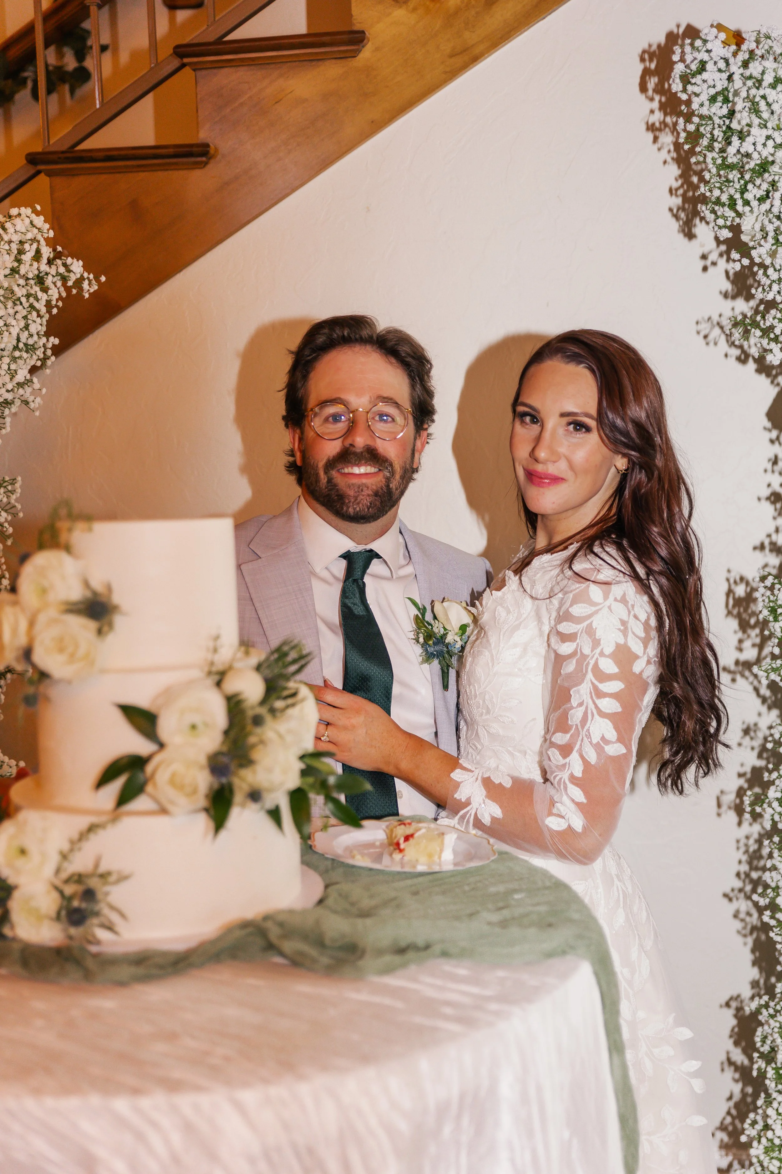 A bride and groom stand next to a wedding cake decorated with white flowers, at their wedding reception.