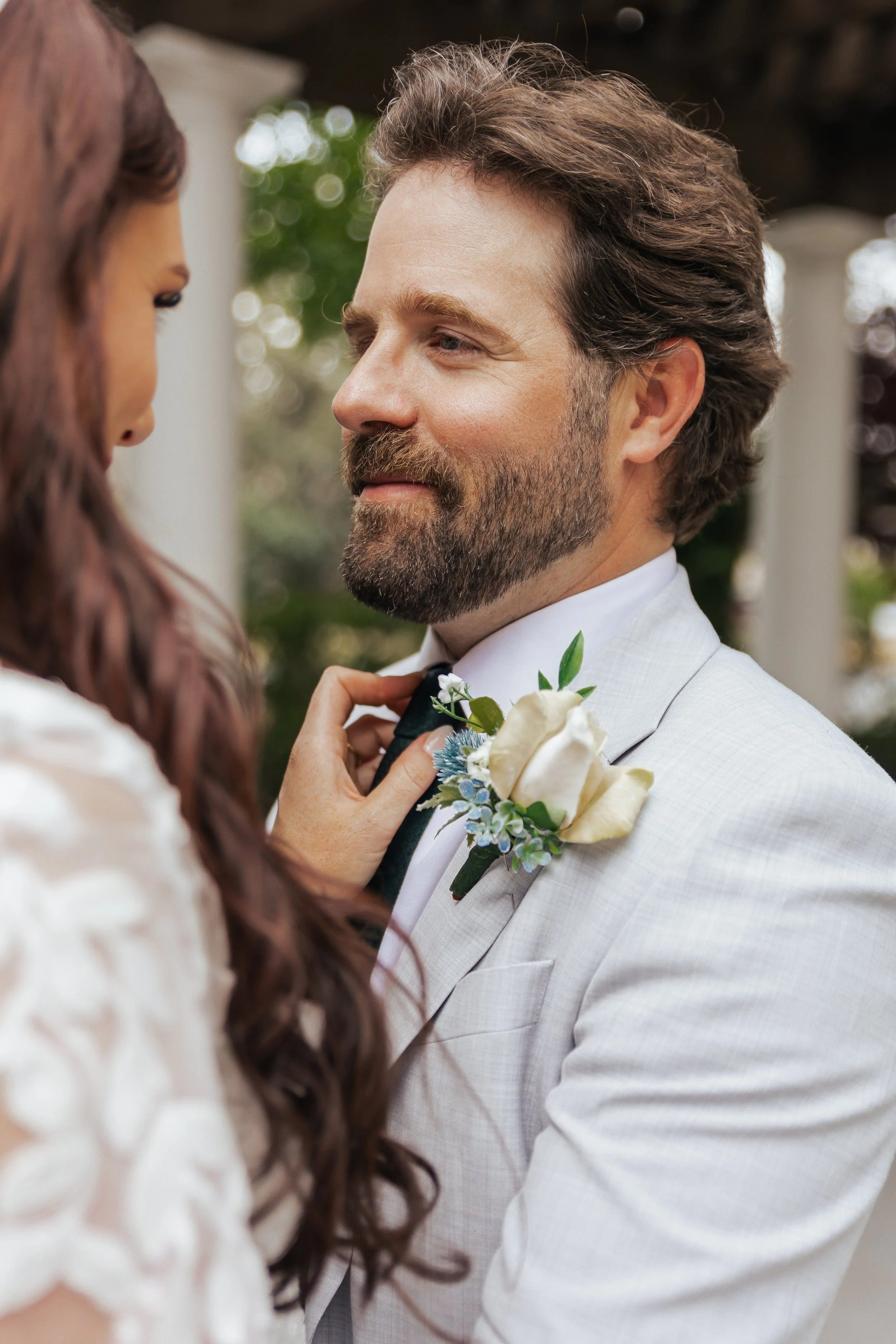 A man in a light gray suit and a boutonniere looks at a woman with long, wavy, reddish-brown hair. The woman, slightly out of focus, adjusts the man's tie. They are outdoors, likely at a wedding or formal event, with blurred greenery in the backgroun