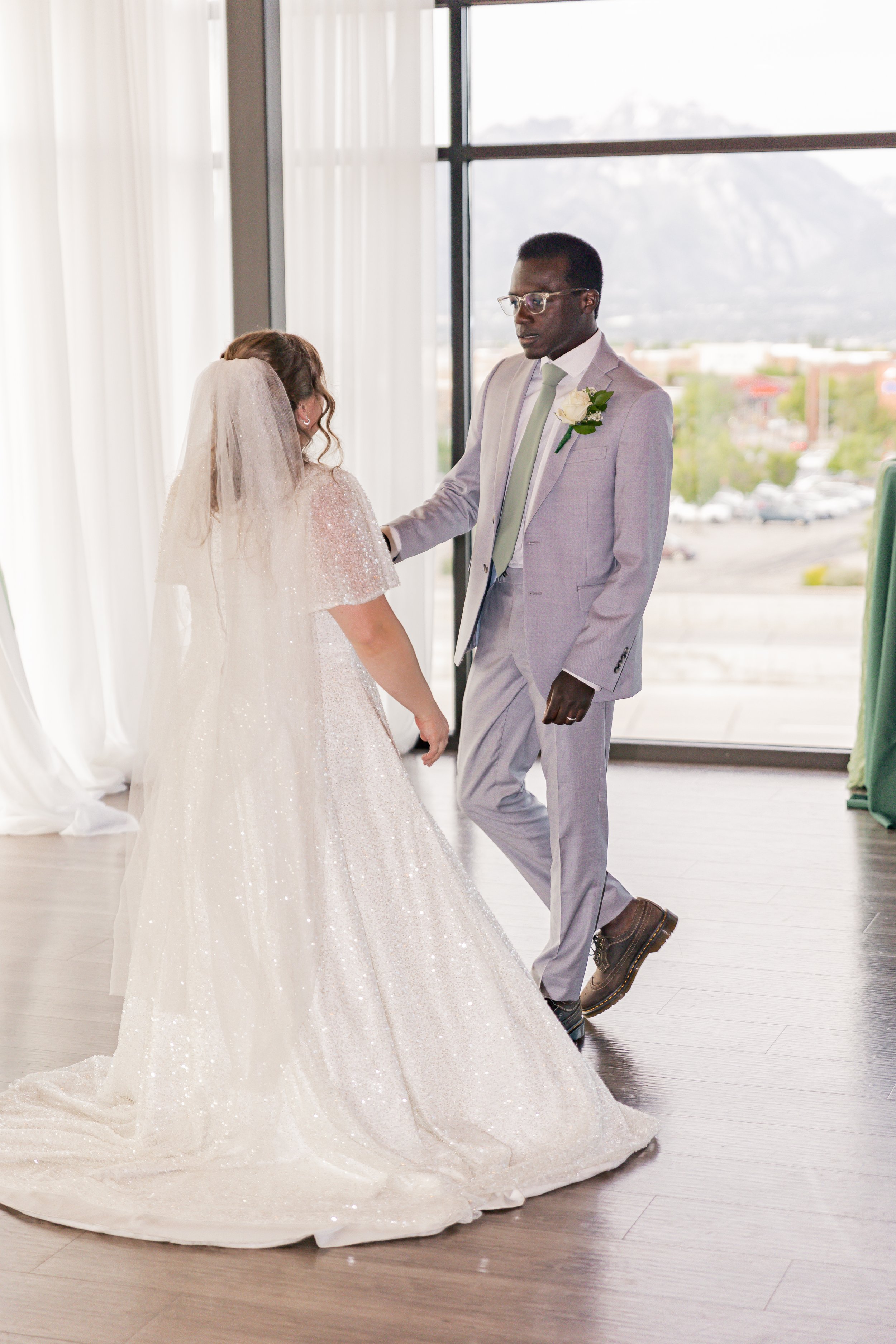 A bride and groom dancing indoors during their wedding, with large windows and a cityscape view in the background.