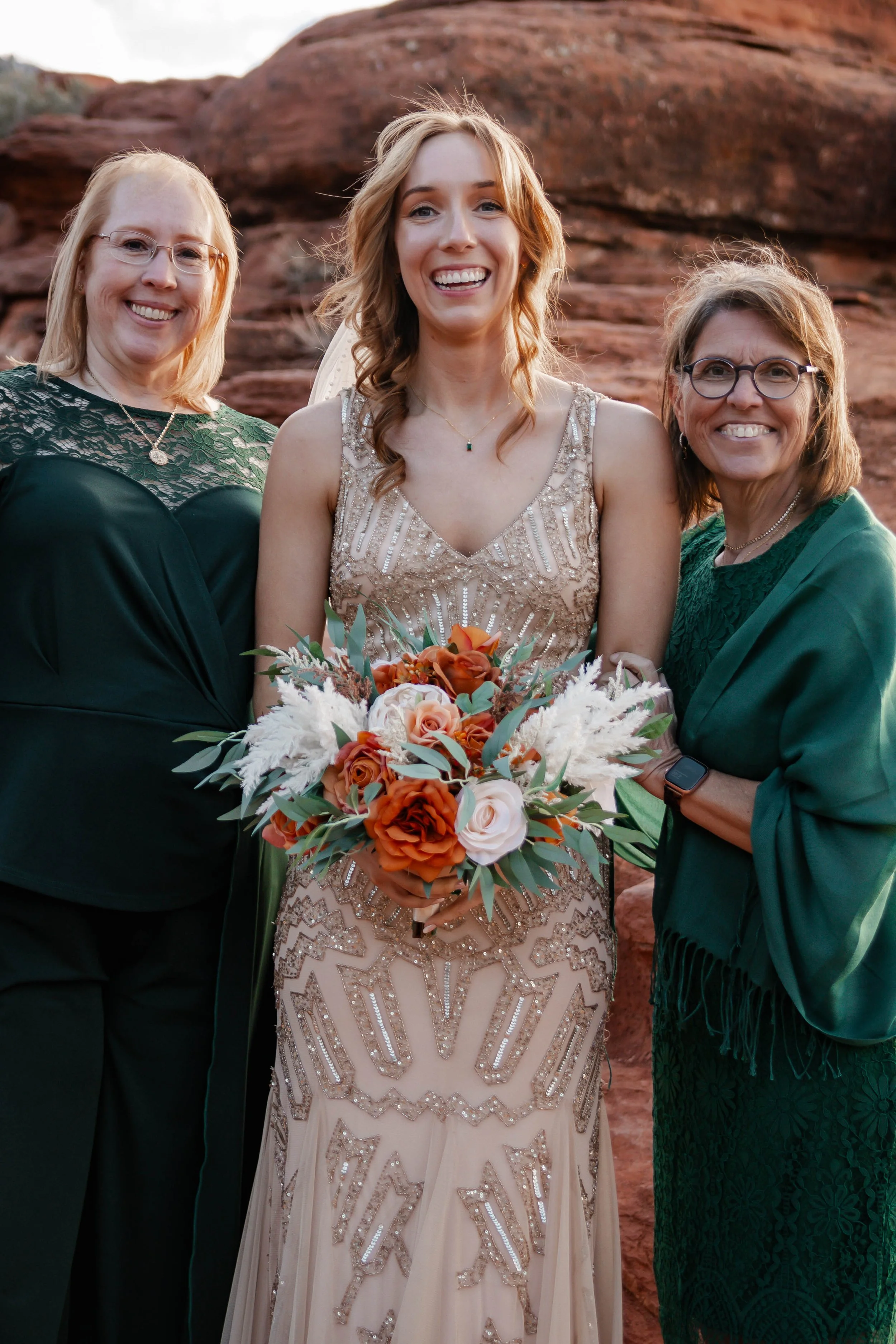 A bride in a beaded gown holding a bouquet of orange, peach, and white flowers, standing with two women in green dresses outdoors with red rock background.