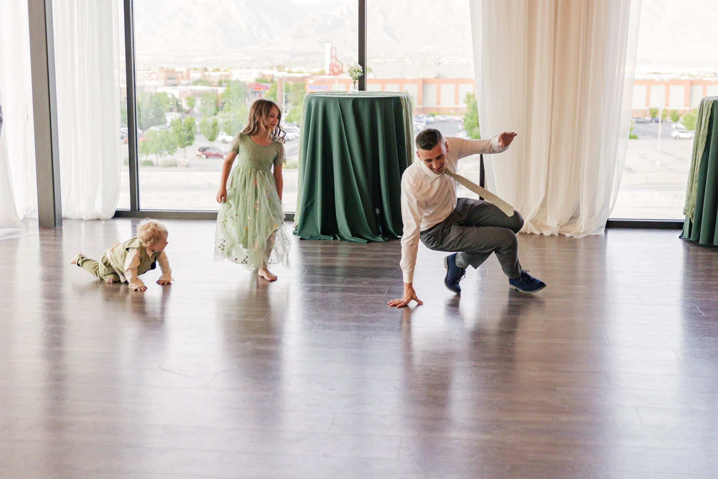 A man in a white shirt and gray pants dancing on a wooden floor with three children, one crawling, one walking, and one standing, in front of large windows with white curtains in a bright room.