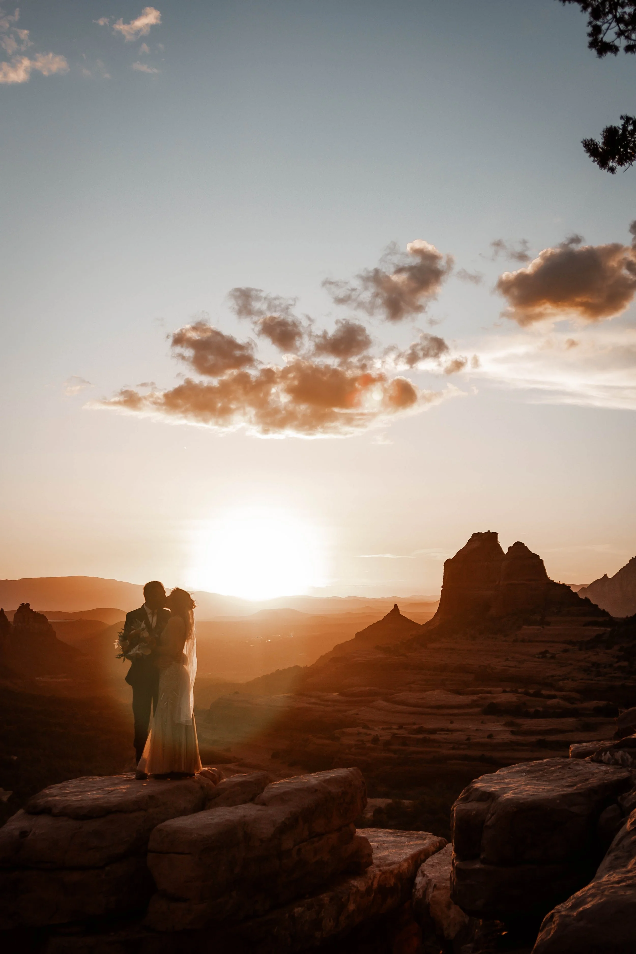 A couple appears to be kissing or embracing on rocks during a sunset in a desert landscape with large rock formations and a few clouds in the sky.