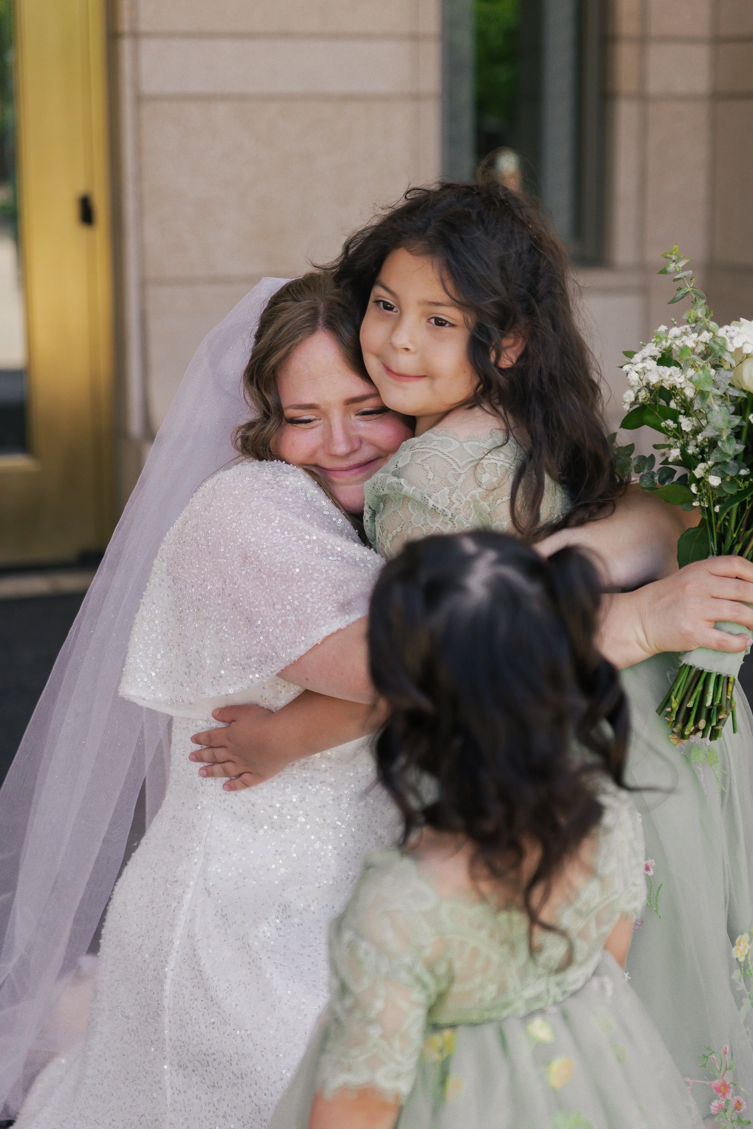 A bride in a white wedding dress hugs a young girl with long black curly hair, who is holding a bouquet of white flowers. Another small girl with dark hair in pigtails, wearing a gray dress with floral embroidery, is looking at the bride. They are sh