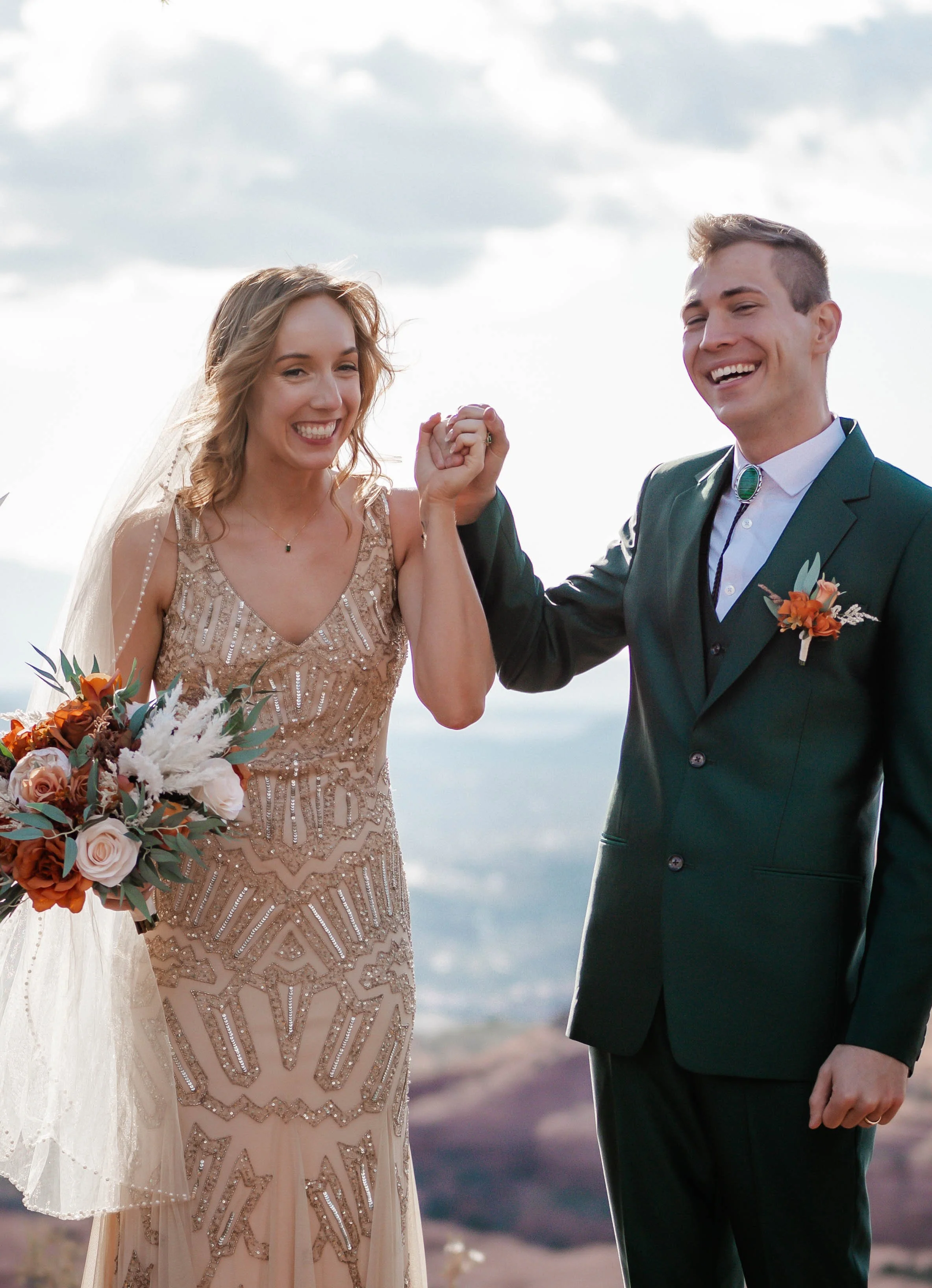 A bride and groom holding hands and smiling outdoors with mountains and sky in the background.