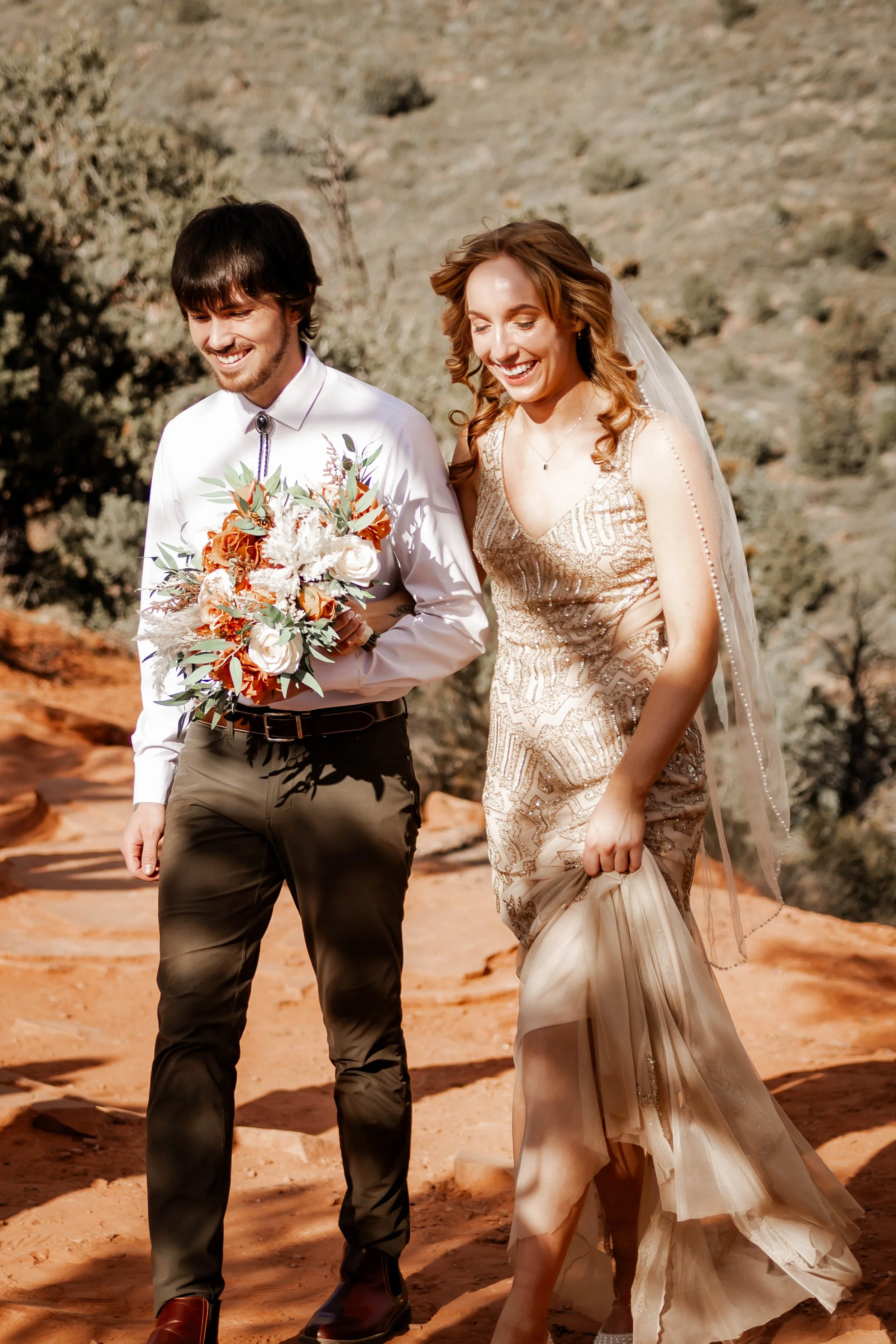 A bride and groom walking outdoors on a sunny day, smiling, with the bride holding a beige dress with layers and the groom holding a bouquet of flowers.