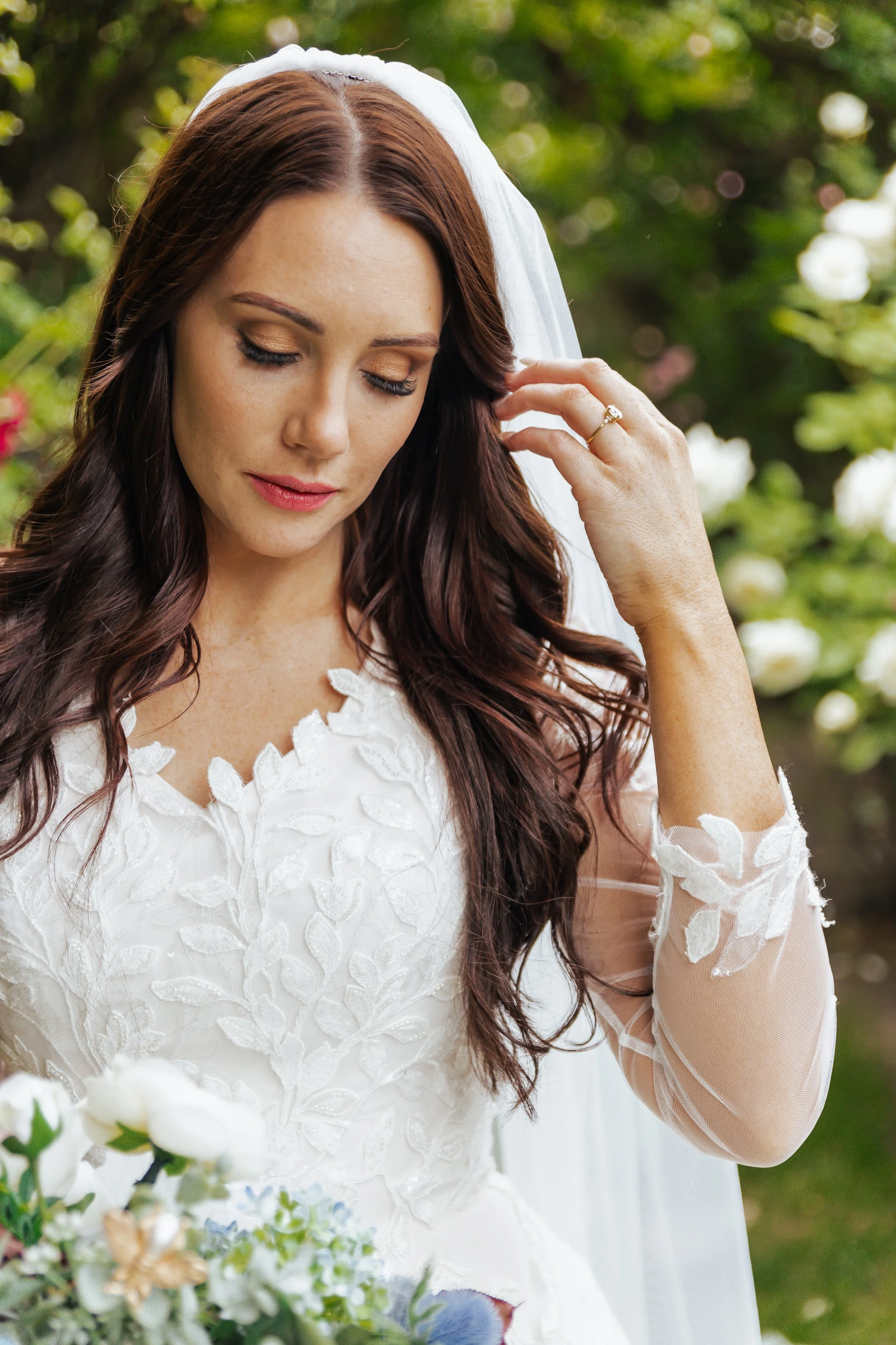 Close-up of a bride with long brown hair wearing a white lace wedding dress and veil, holding her hair, outdoors with green foliage in background.