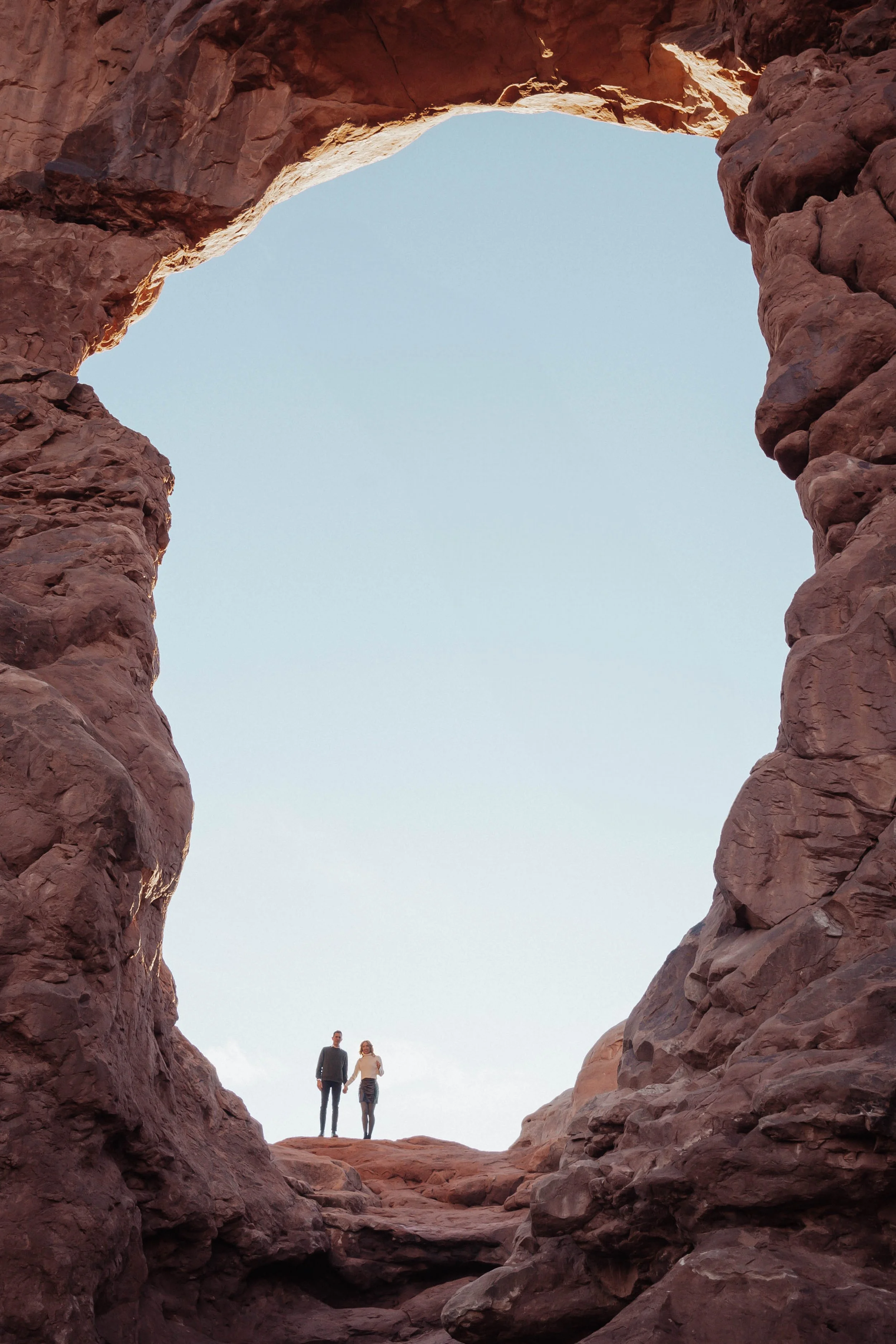 Two people holding hands standing inside a natural rock arch formation in Arches National Park with a clear sky background.