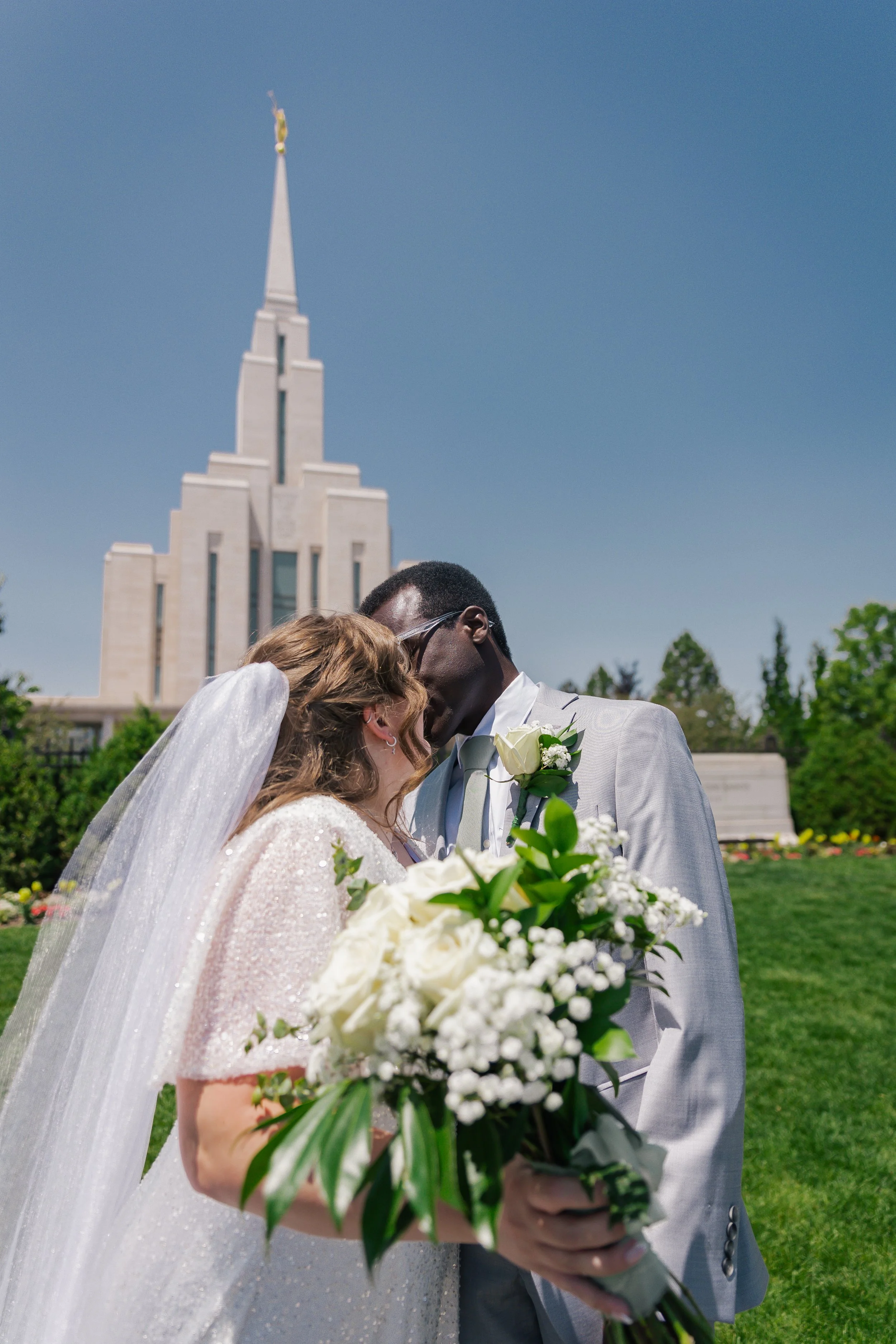 A bride and groom are close together outdoors, sharing a kiss, with the bride holding a bouquet of white flowers, with oquirrh mountain temple background.