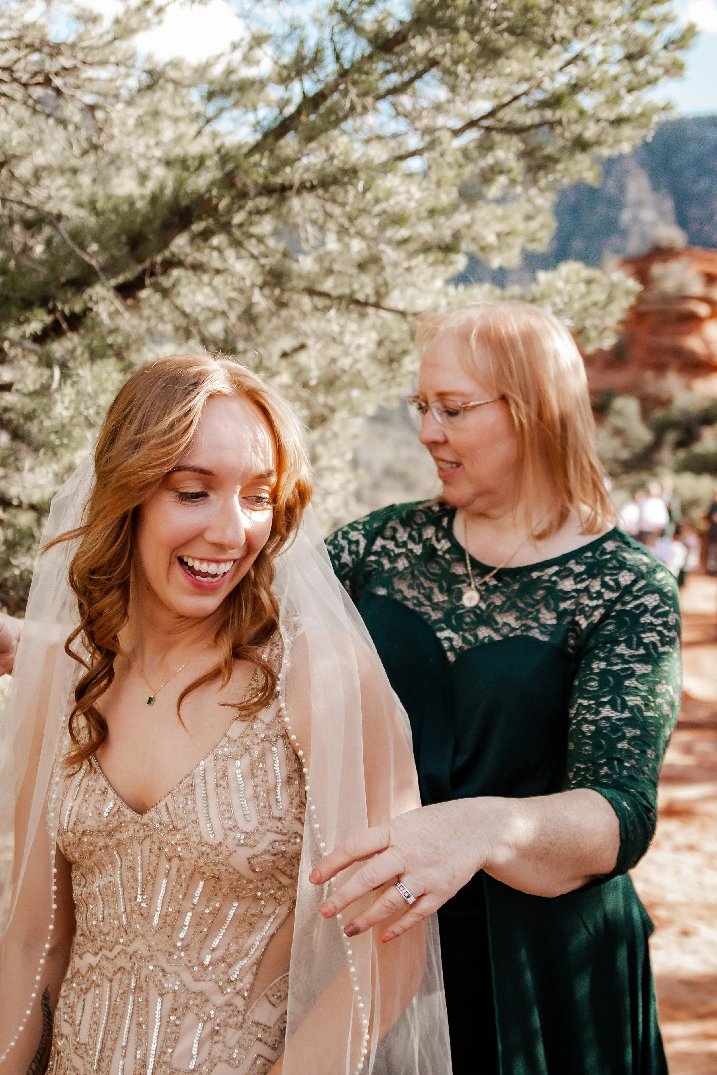 A bride in a beige dress with beadwork and a veil smiling as she is assisted by a woman in a dark green lace dress with glasses, in an outdoor setting with trees and red rocks in the background.