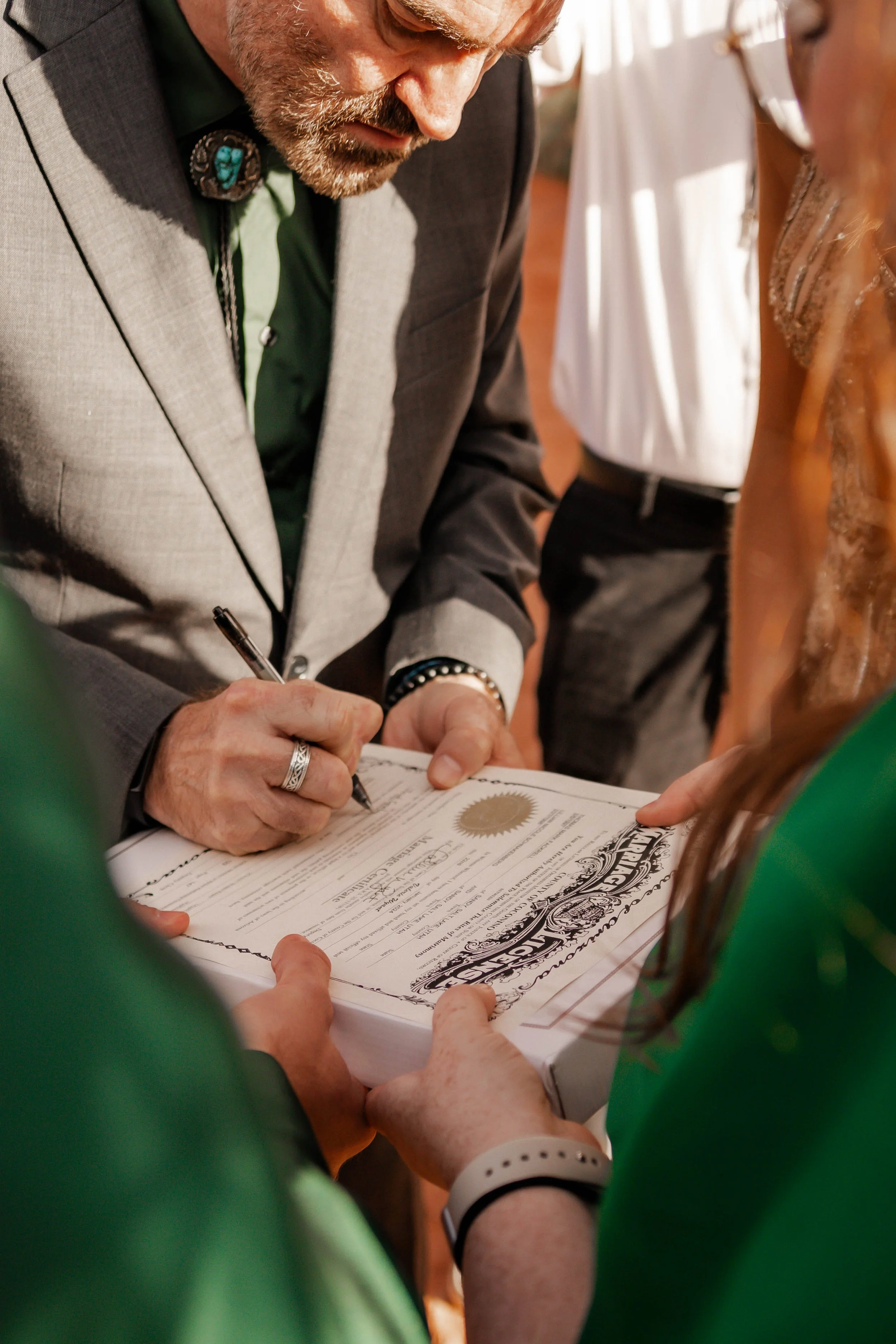 A man with gray hair and beard signing a legal document or certificate, surrounded by others at a formal event.
