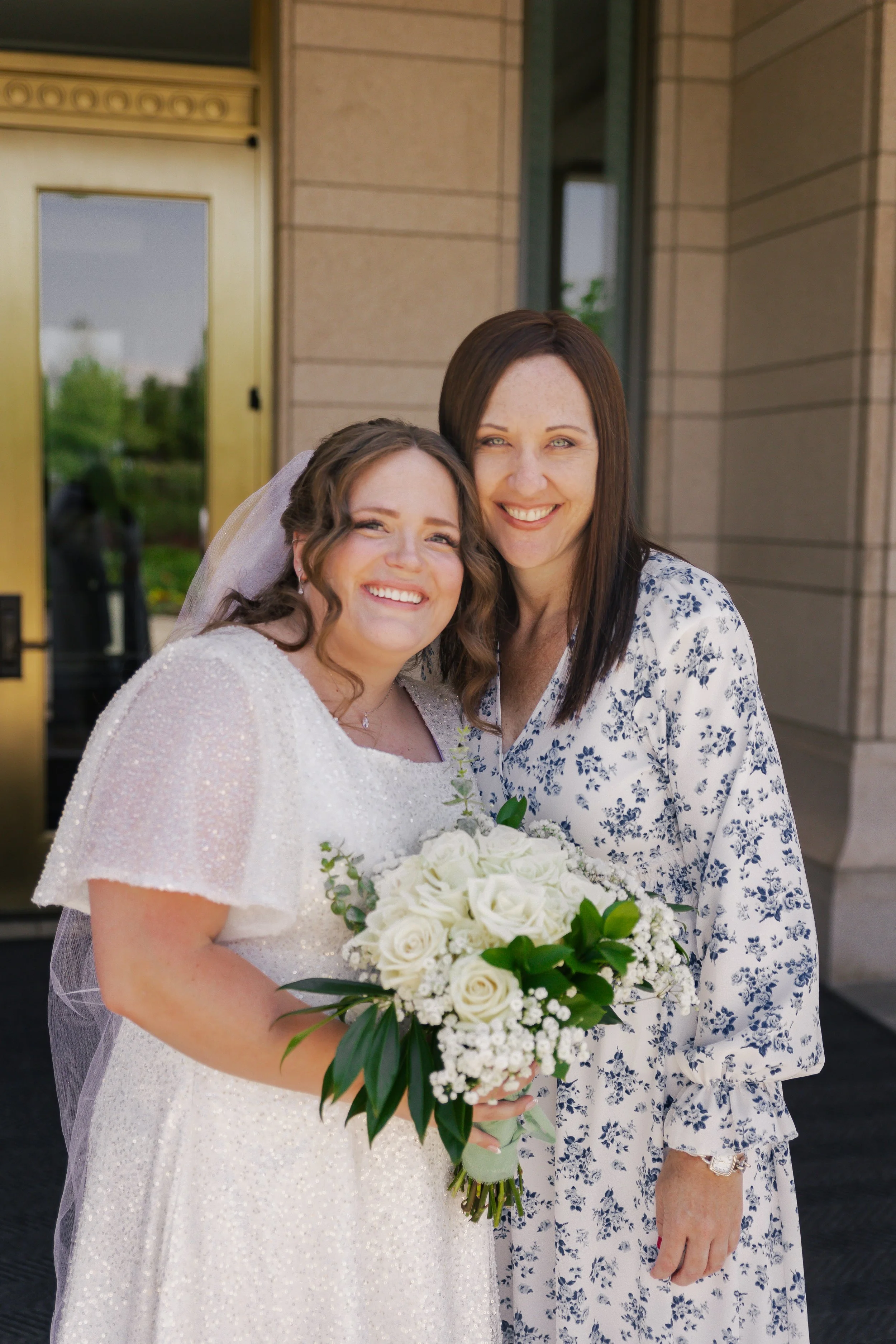 A bride holding a bouquet of white roses and greenery, smiling with a woman in a floral dress outside a building.
