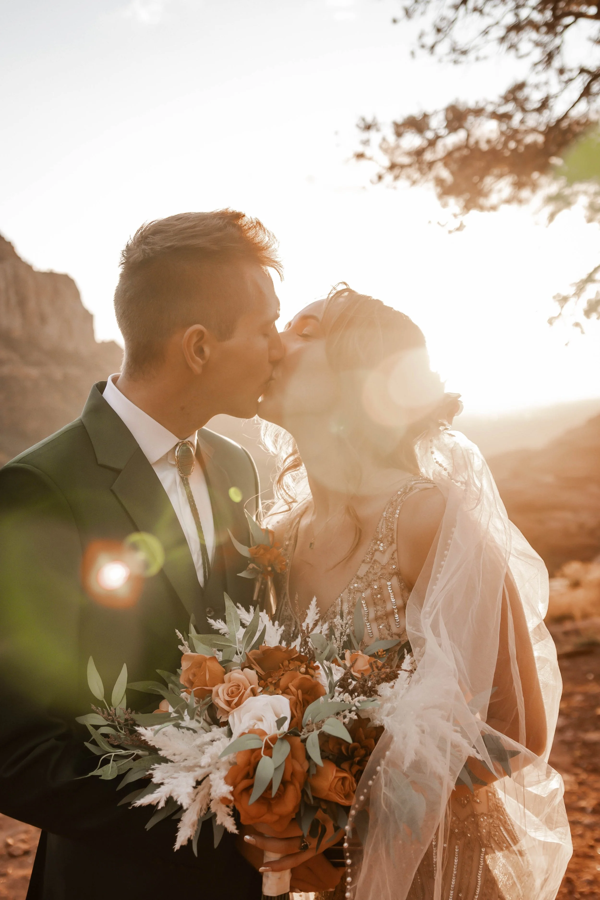 A couple dressed in wedding attire sharing a kiss outdoors during sunset. The groom wears a dark suit with a bolo tie, and the bride wears a detailed dress with a veil, holding a bouquet of orange and beige flowers with greenery.