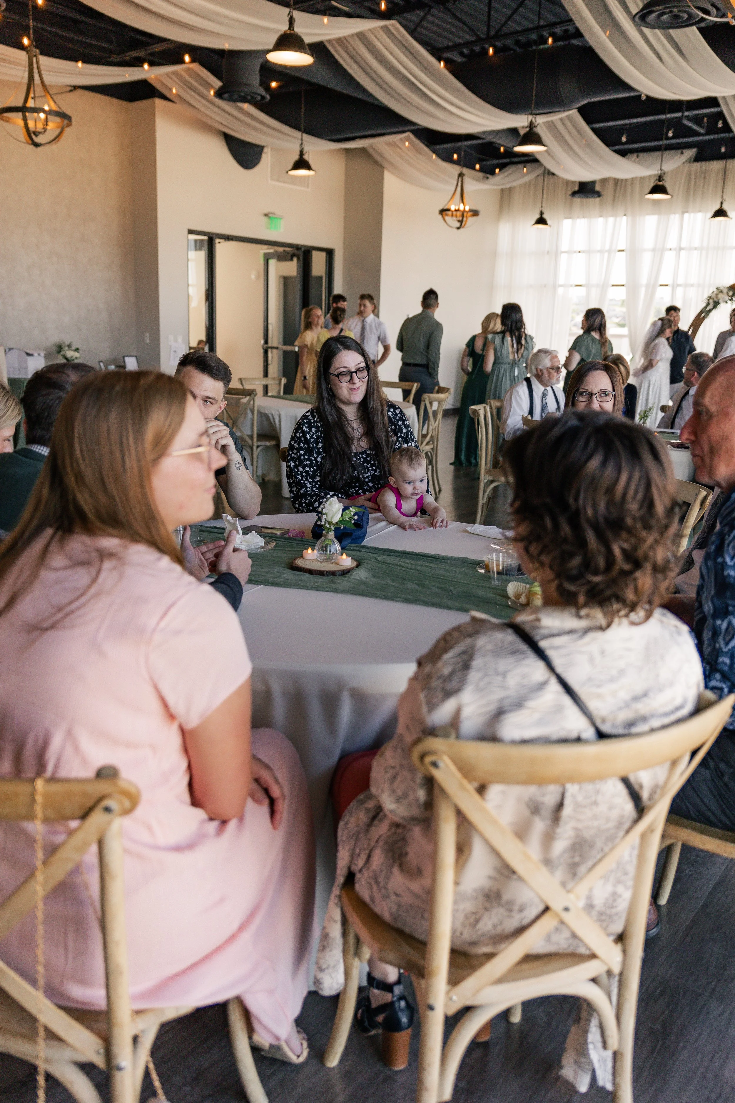 People sitting around a decorated banquet table at an indoor event, with some guests standing in the background, under draped fabric and ceiling lights.