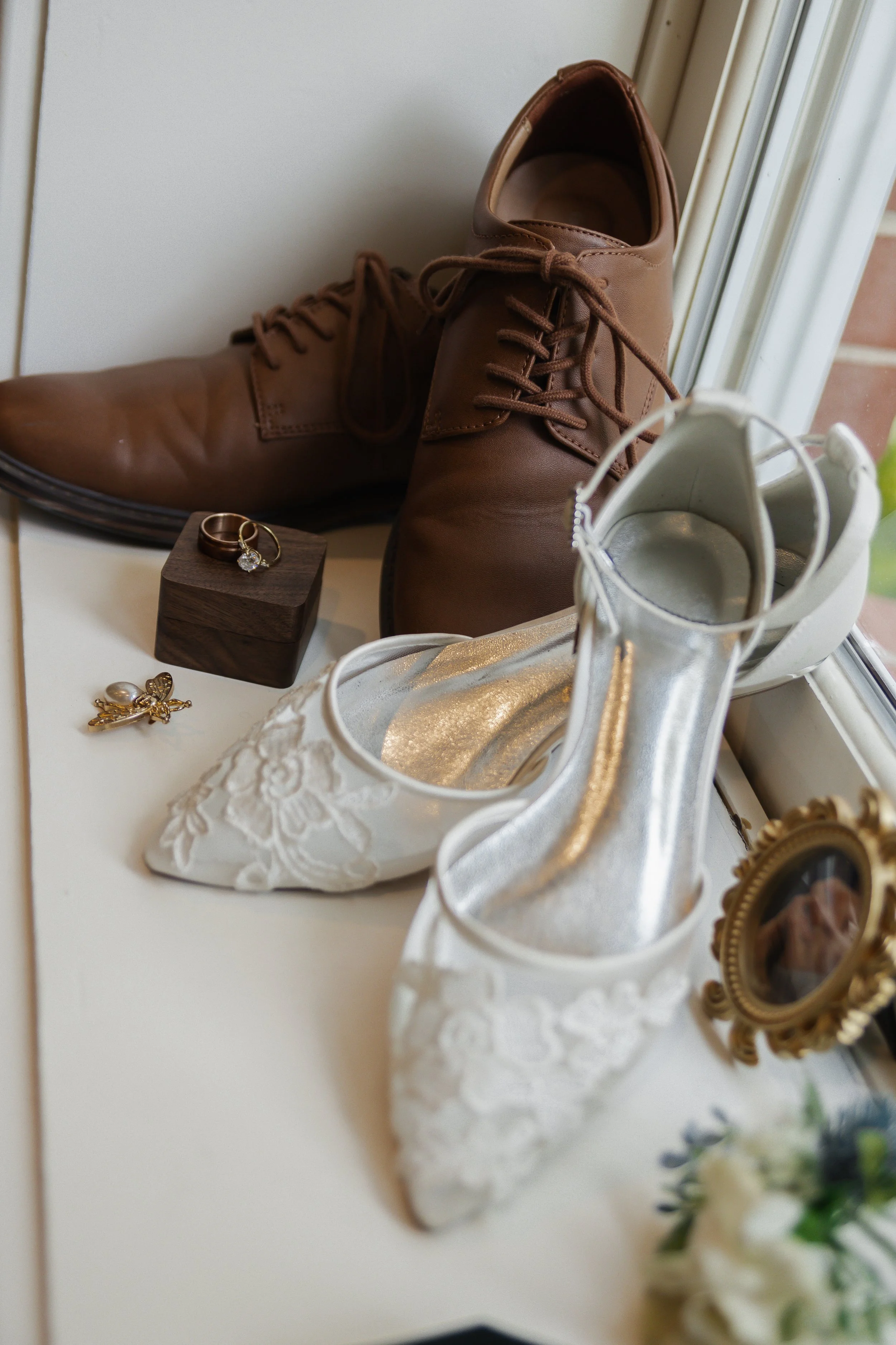 Wedding shoes and accessories on a windowsill, including white lace wedding shoes, brown dress shoes, a ring box with a ring, and jewelry.