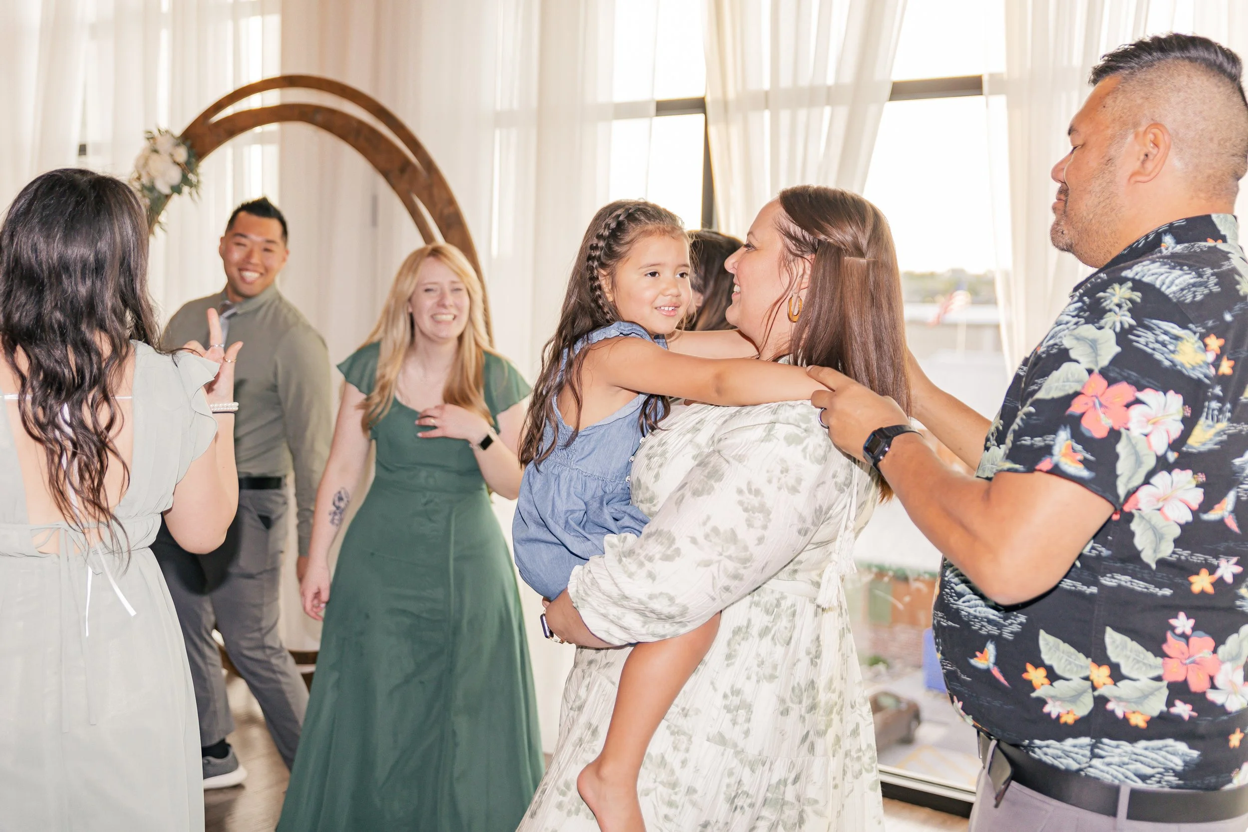 Group of people celebrating, with a woman holding a young girl who is smiling, and others clapping and laughing in the background, during a joyful occasion indoors.