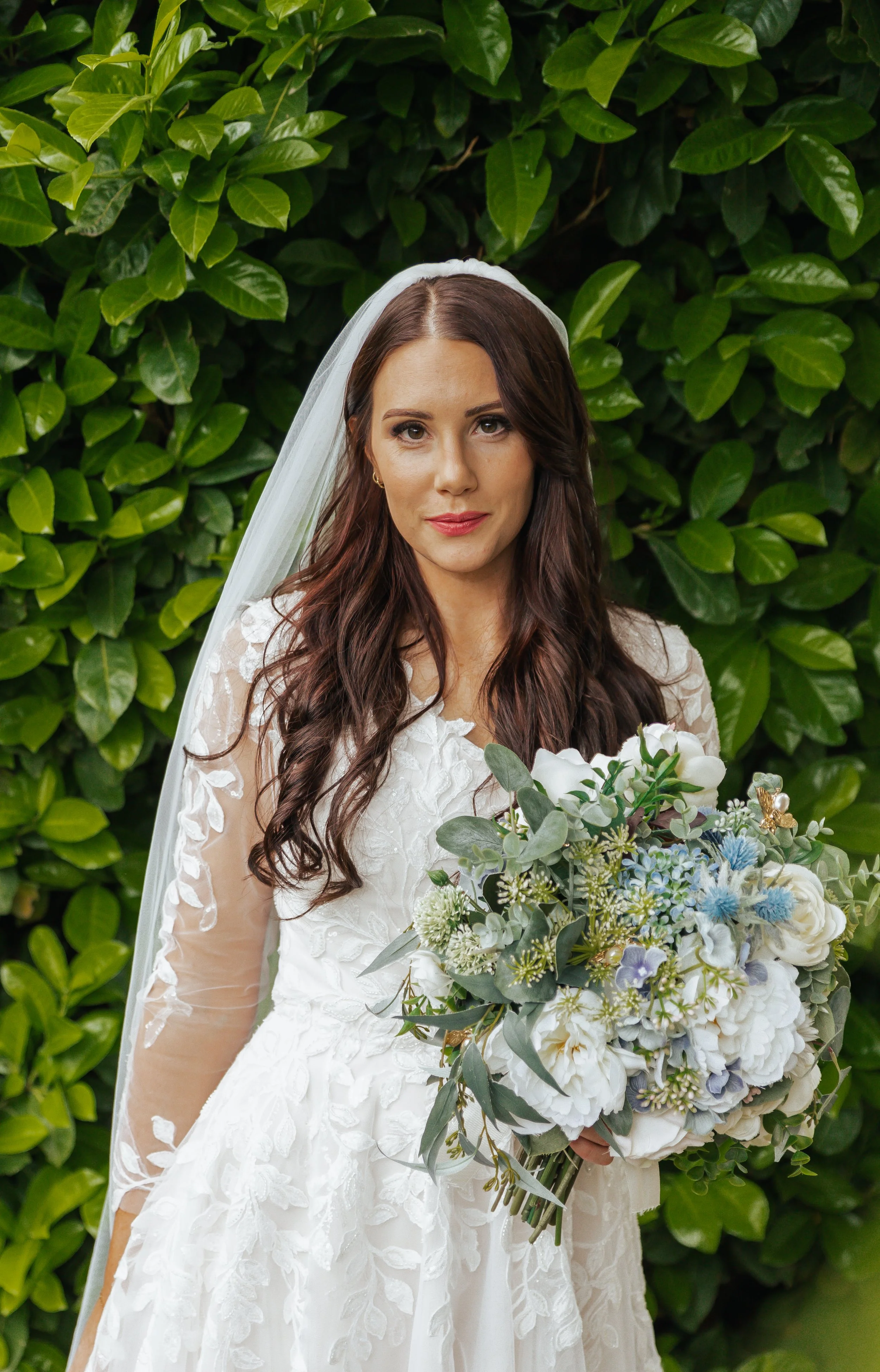A bride in a white wedding dress with lace details, holding a large bouquet of white and blue flowers, standing in front of a green leafy background.