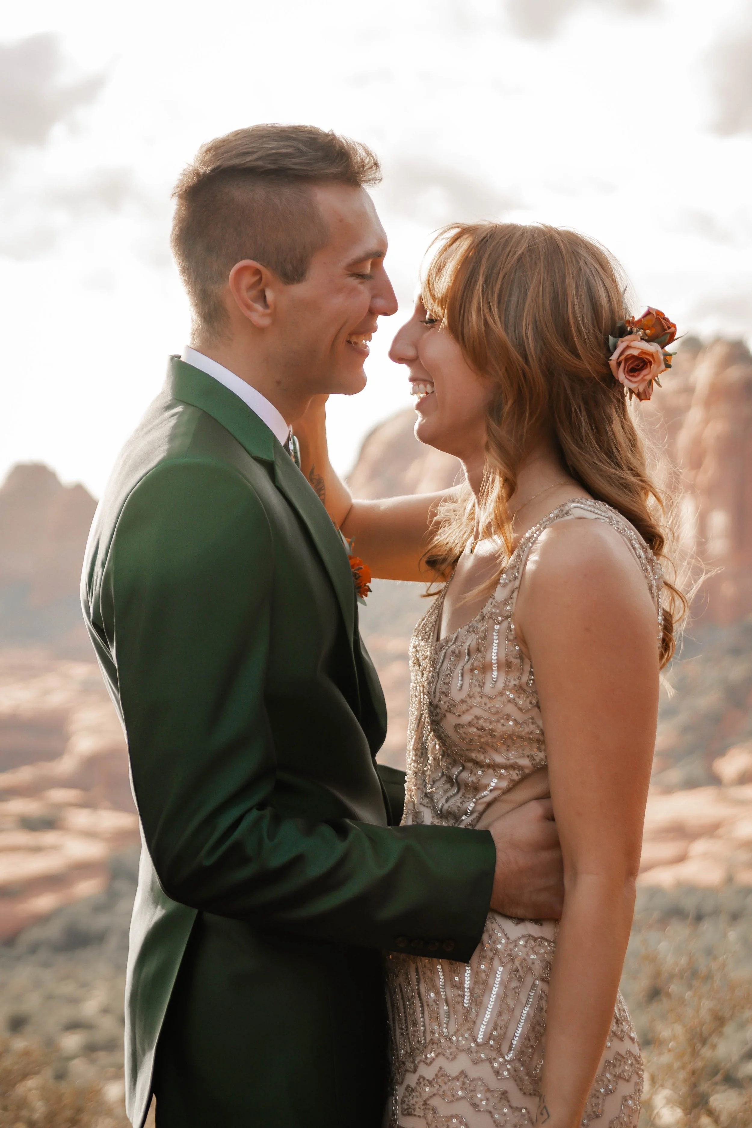 A couple dressed in wedding attire, smiling and touching foreheads, outdoors with a rocky desert landscape and cloudy sky in the background.