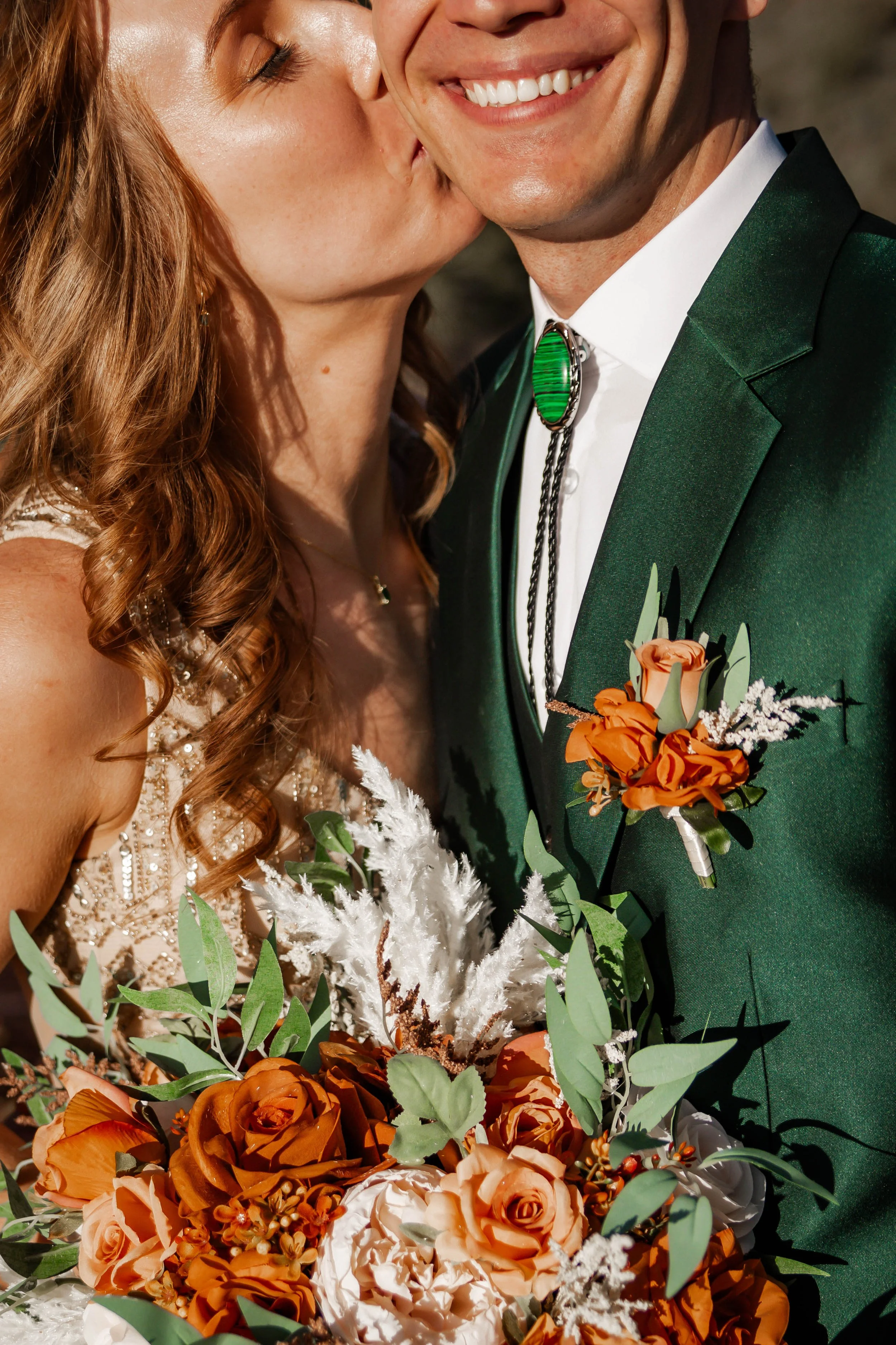 A close-up of a bride and groom at their wedding, with the bride kissing the groom on the cheek, both smiling. The bride has long curly red hair and is wearing a white dress with lace details, holding a vibrant bouquet of orange roses, white flowers,