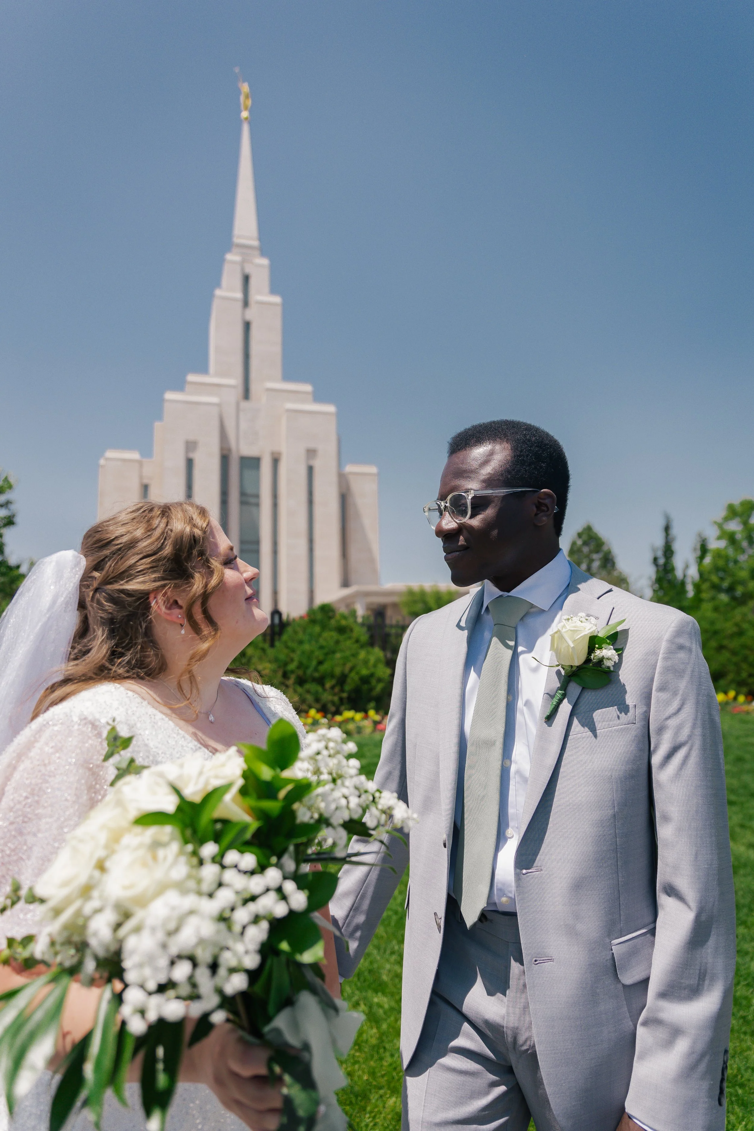Bride and groom standing outdoors in front of oquirrh mountain temple, sharing a moment on their wedding day, with the bride holding a bouquet of white flowers.