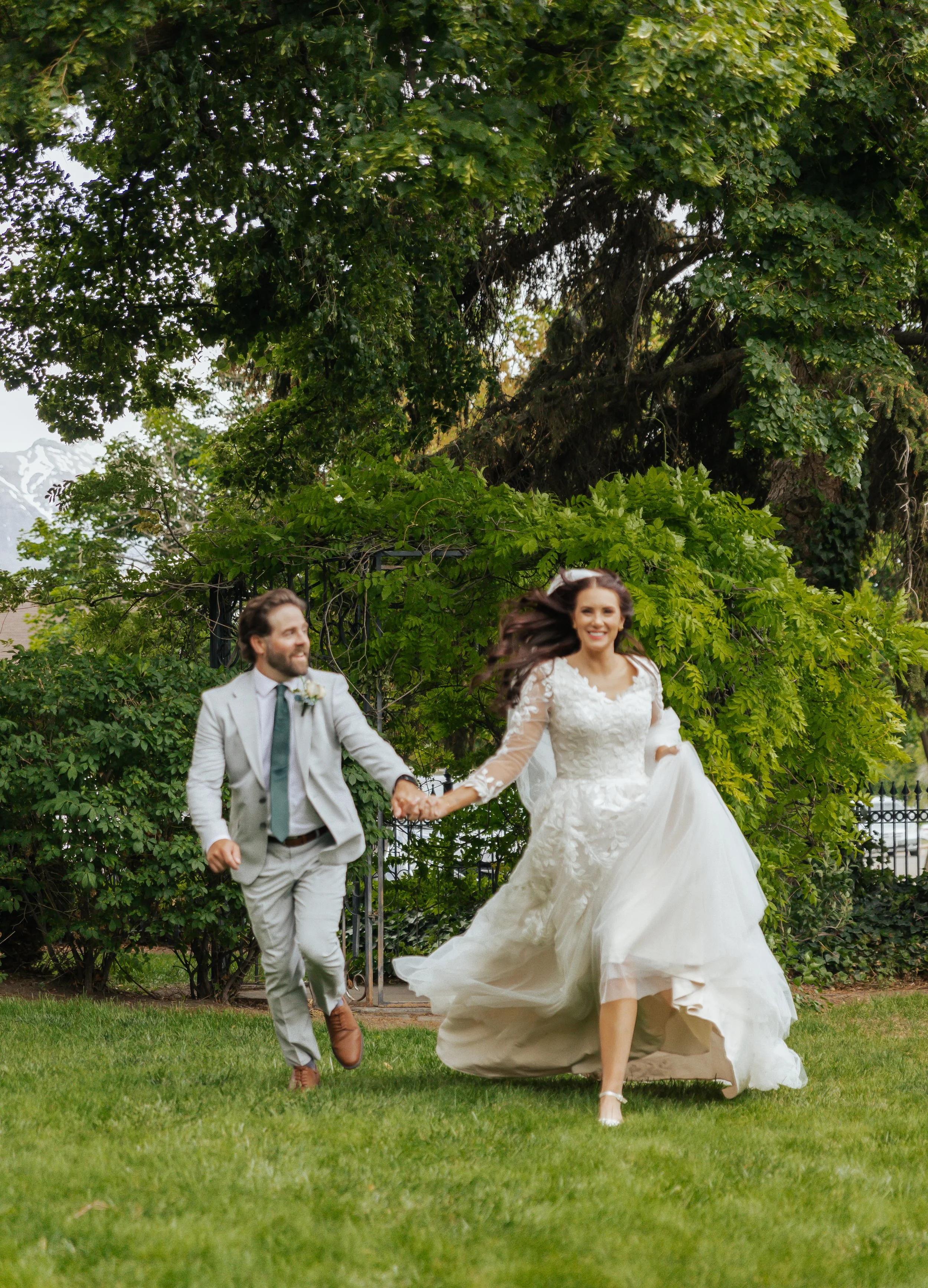 A bride and groom holding hands and running through a lush green garden on their wedding day, with trees and a fence in the background.