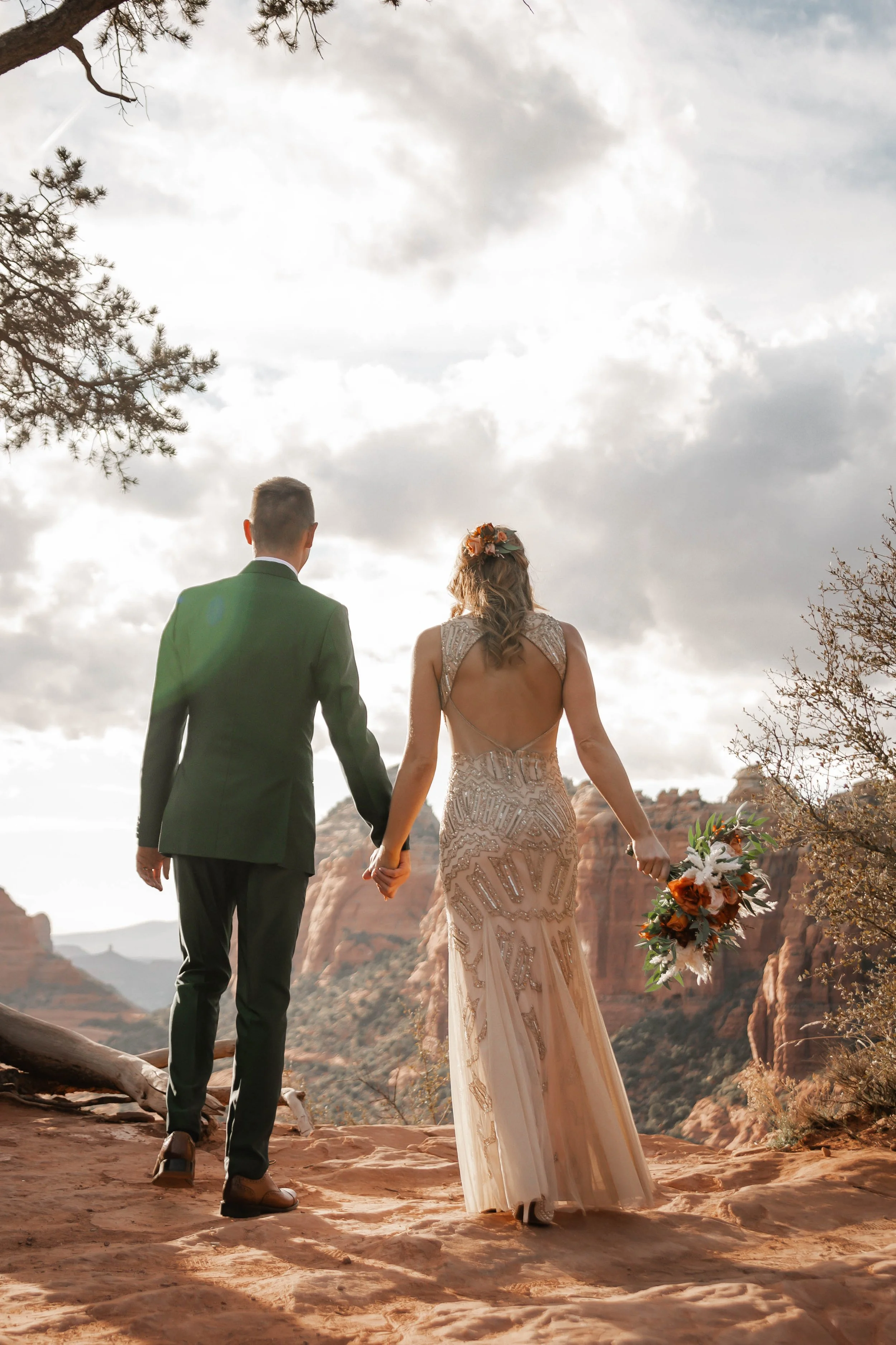 A bride and groom holding hands walking in a desert-like landscape with red rock formations and a cloudy sky.