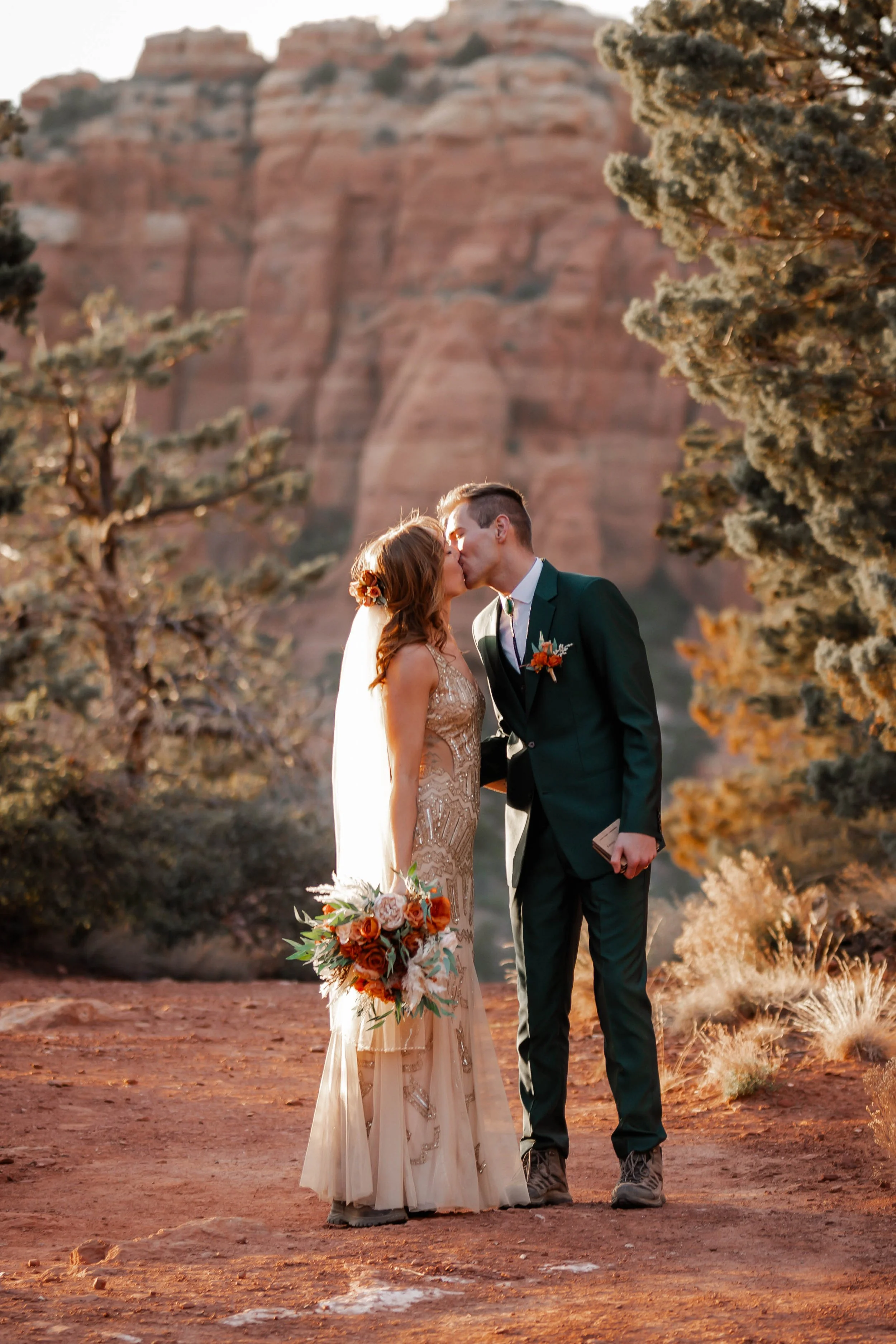 A couple in wedding attire sharing a kiss outdoors, surrounded by trees and red rock formations, during sunset.