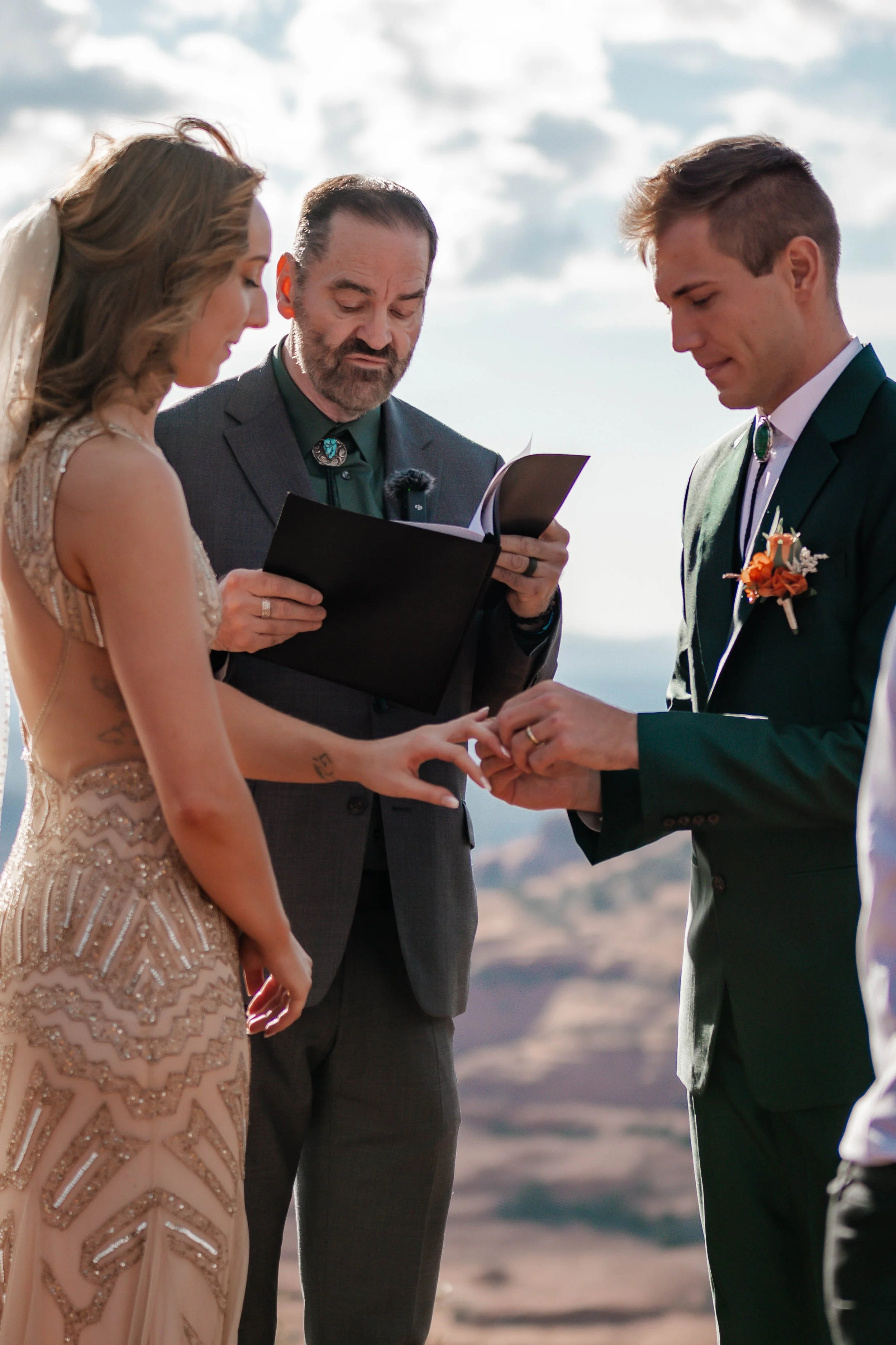 A wedding ceremony outdoors during the daytime with a couple exchanging rings, officiant holding a book, and mountains in the background.