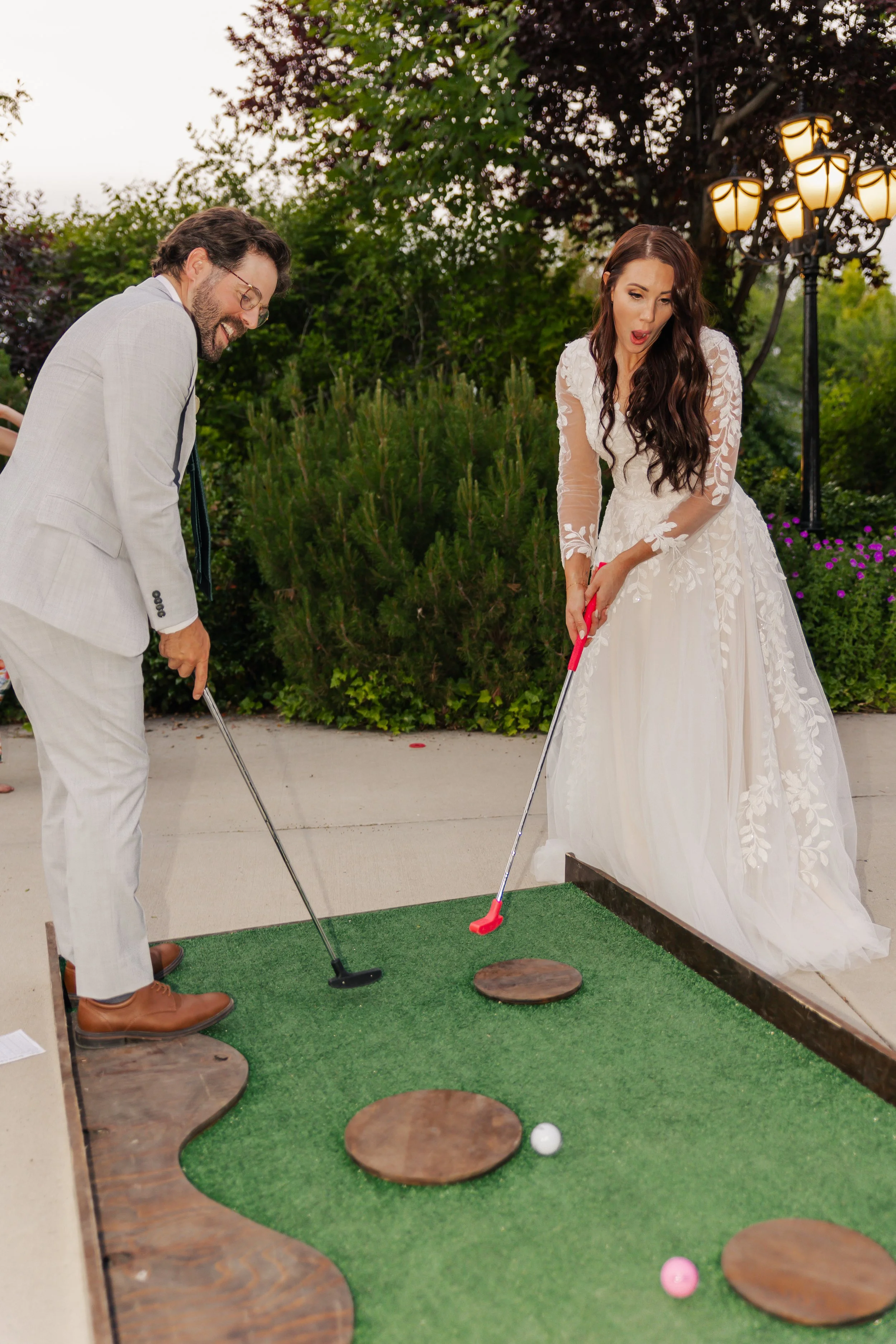 Bride and groom playing mini-golf outdoors on a small green course, dressed in wedding attire.