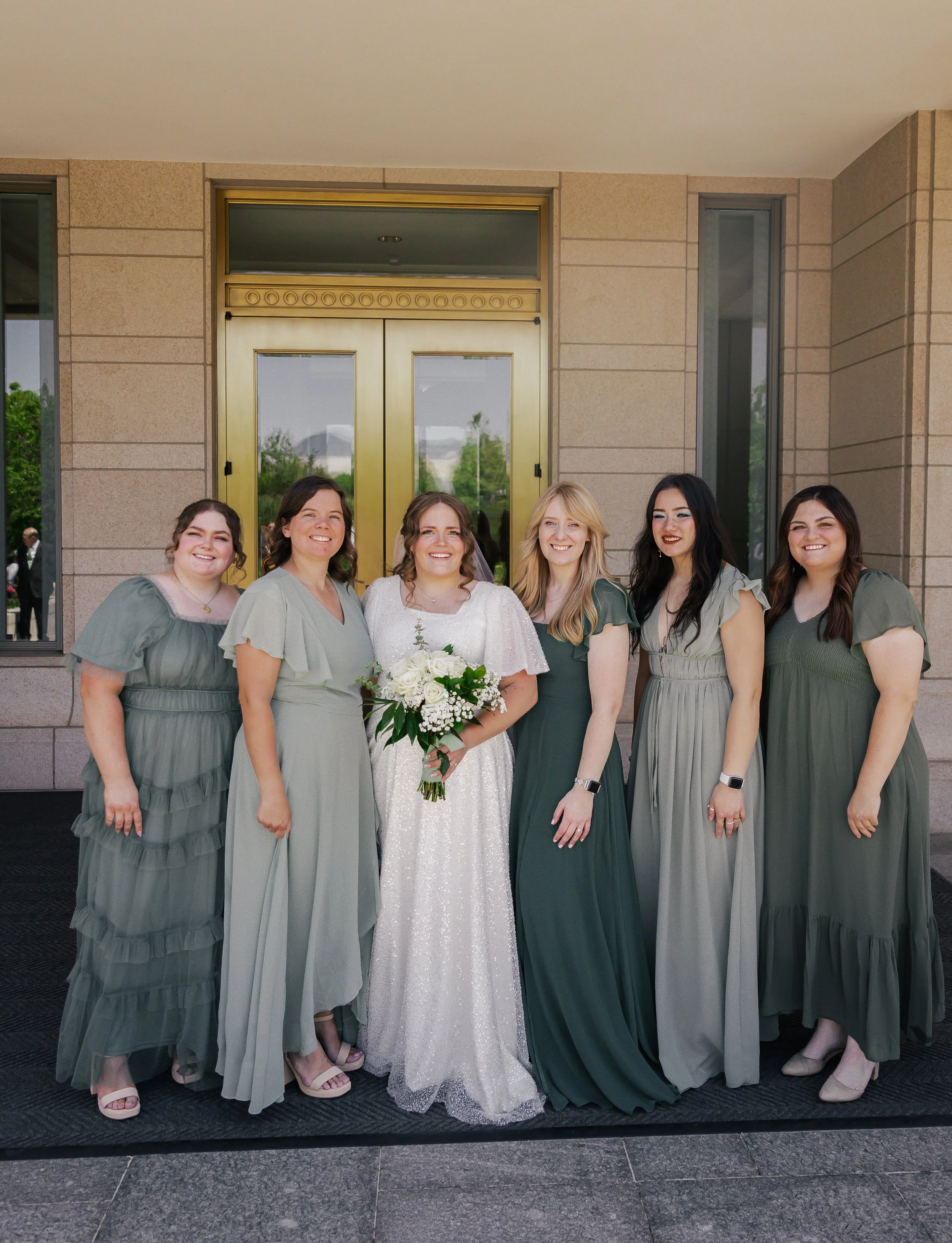 A bride and five bridesmaids standing in front of a building with brick walls and a golden door, smiling for a photo. The bride is holding a bouquet of white flowers.