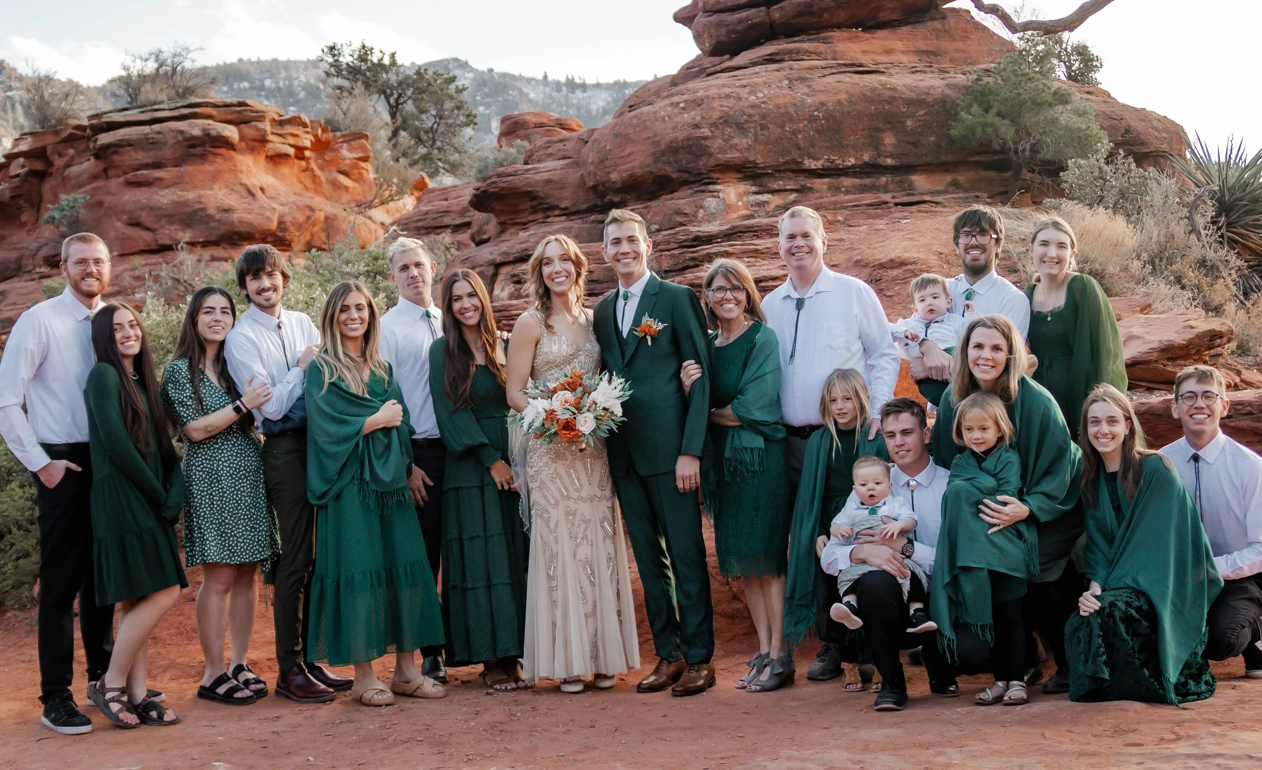 A large group of people in wedding attire, including bride and groom, standing outdoors against red rock formations with dry desert plants in the background.