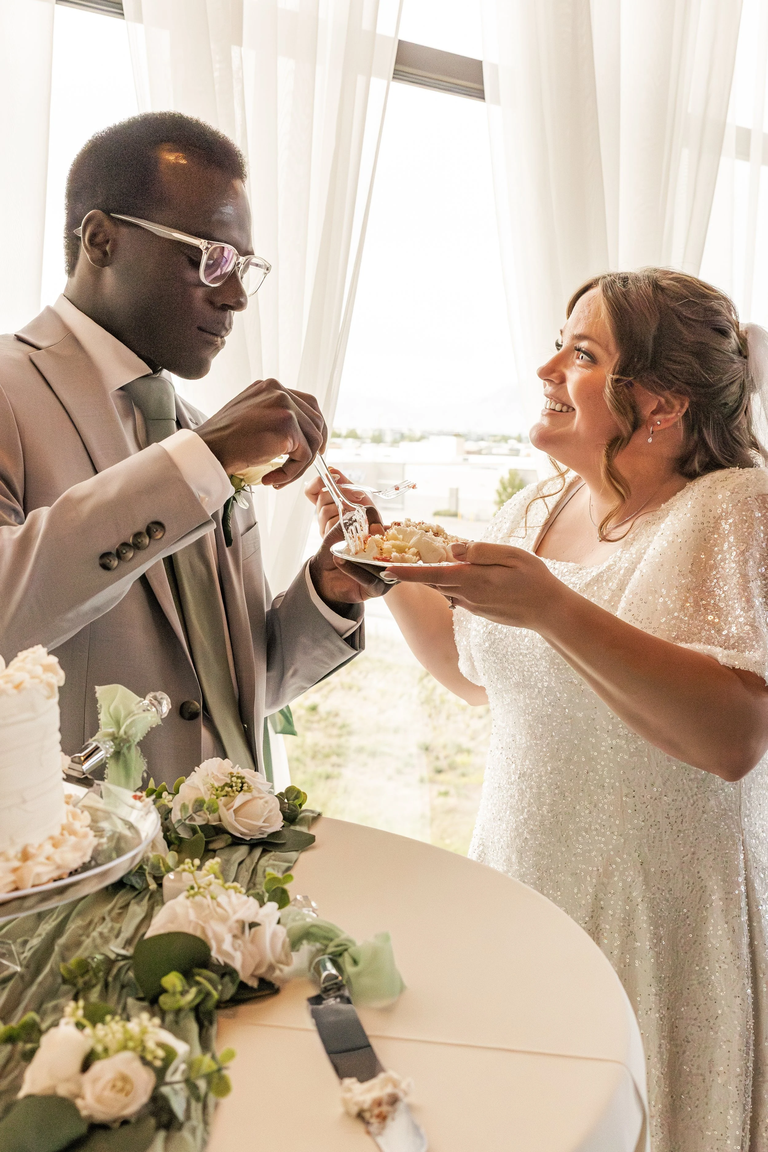 A bride and groom sharing a piece of cake at their wedding reception indoors with sheer curtains and a bright background.