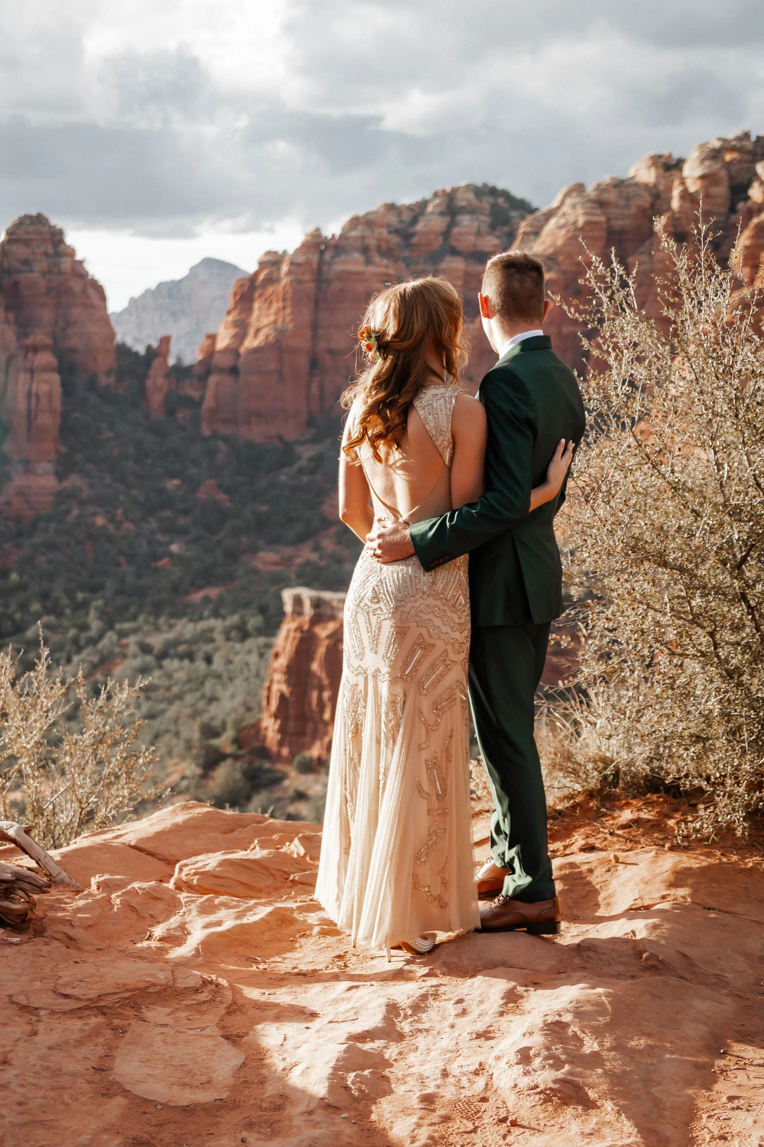 A bride and groom stand close together on a rocky desert landscape with red cliffs in the background, embracing each other during sunset.