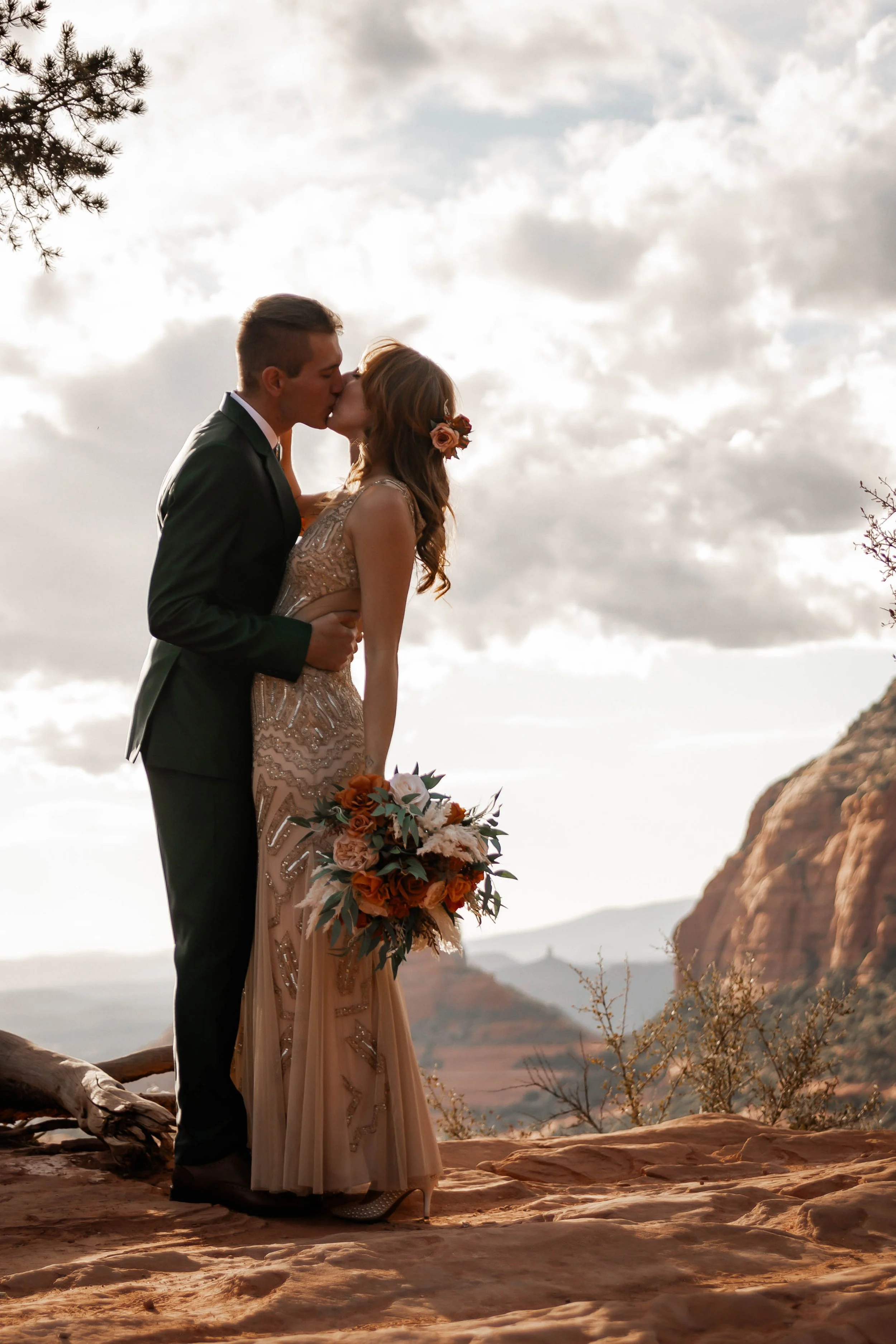 A couple in wedding attire sharing a kiss outdoors on a rocky landscape with mountains in the background during sunset.