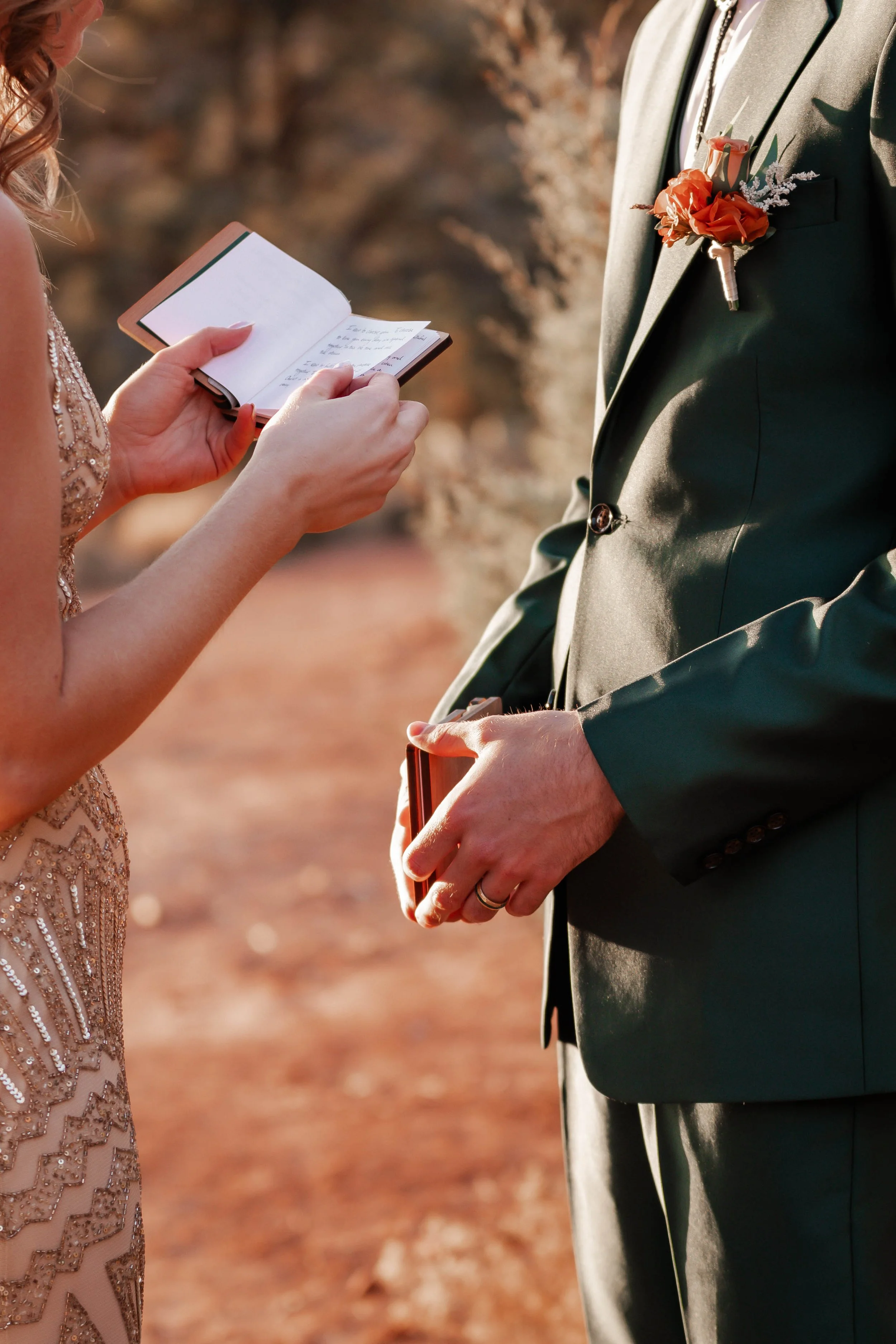 A woman is reading vows from a small notebook during a wedding ceremony with a man dressed in a green suit with a boutonniere of orange and white flowers.