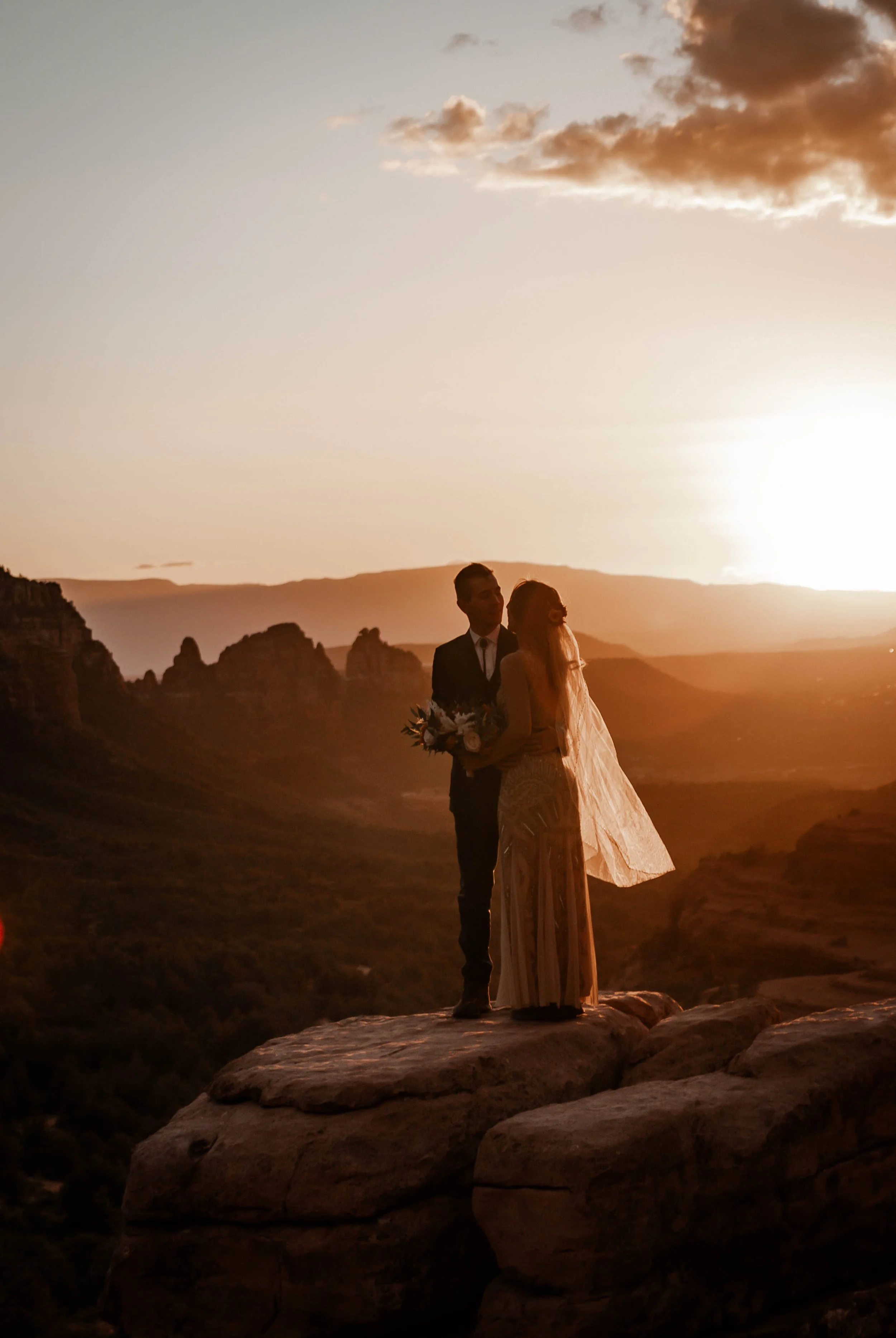 A couple stands on a rock ledge during sunset, with a scenic canyon landscape in the background.