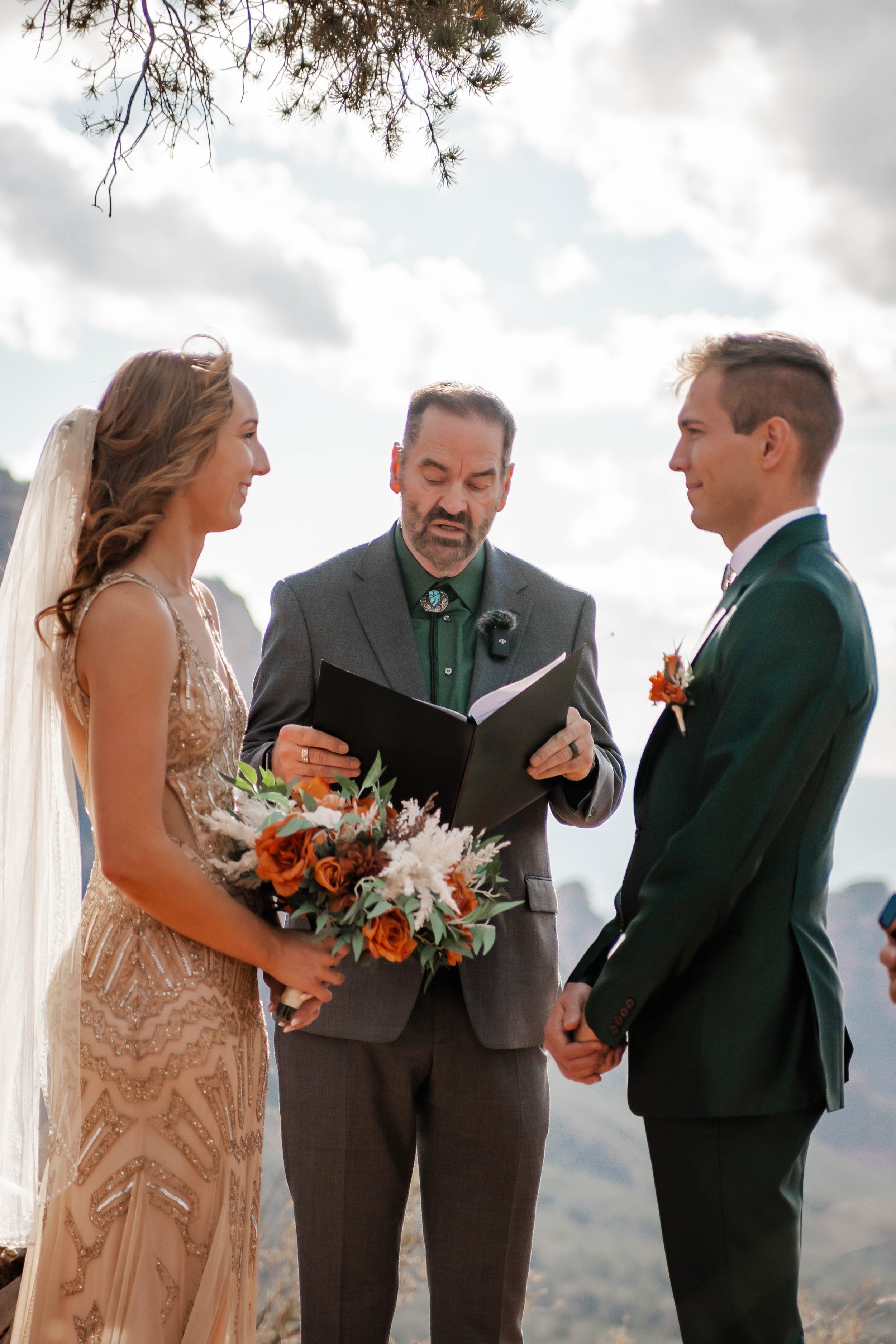 A wedding ceremony outdoors with a bride in a beige dress holding a bouquet of flowers, a groom in a dark green suit, and an officiant reading from a book, with a scenic mountain background and cloudy sky.
