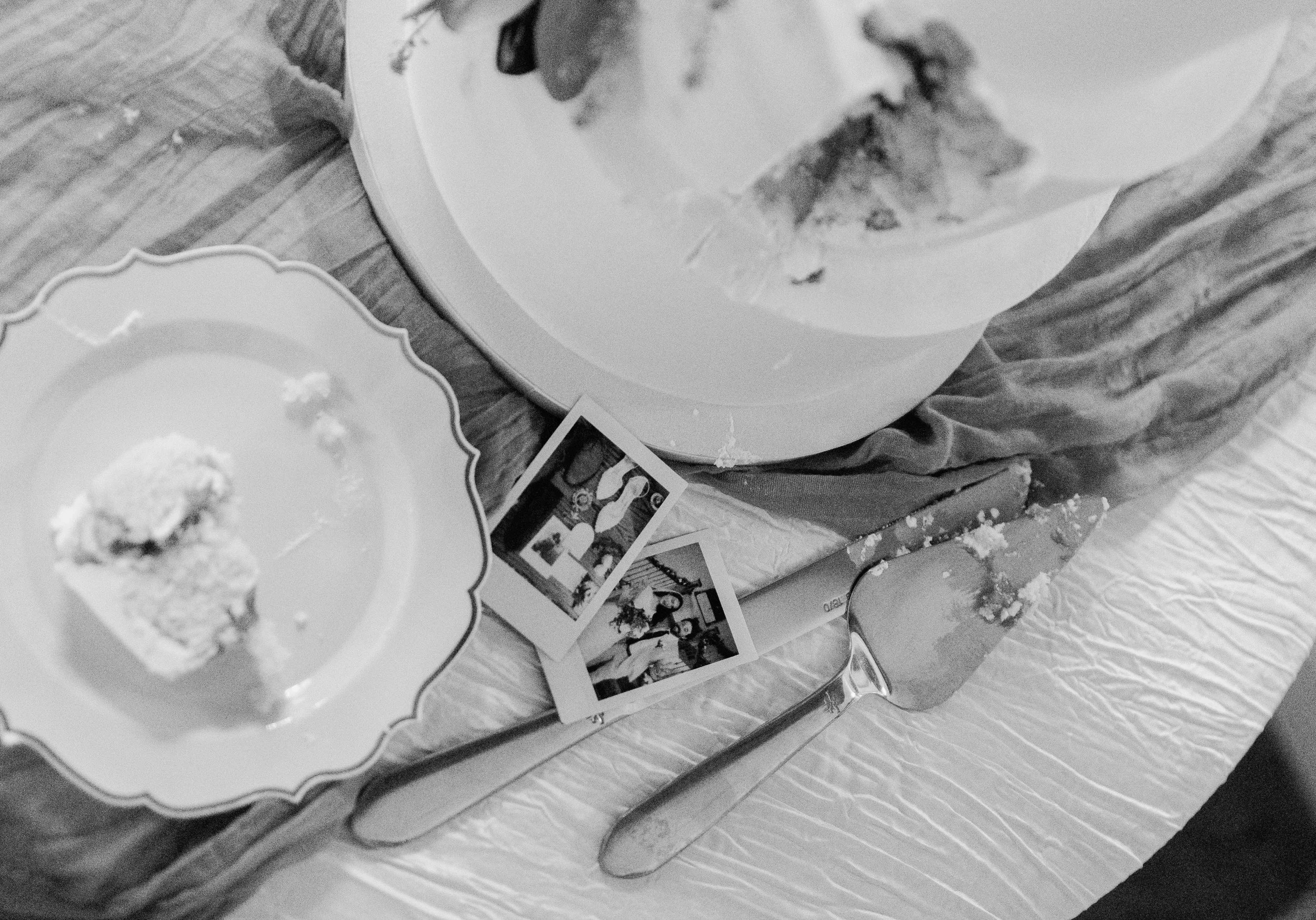Black and white photo of an empty cake plate, a cake server with cake crumbs, and two instant photos on a table with a tablecloth.