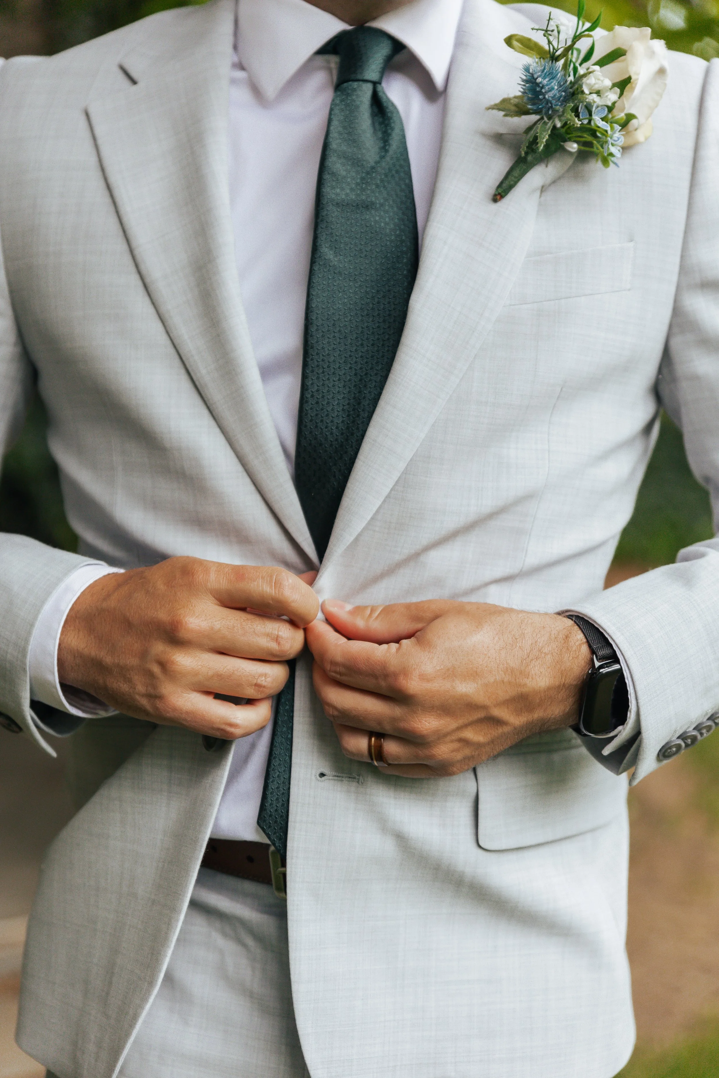 Man in a light-colored suit adjusting his jacket, with a boutonniere on his lapel and a watch on his wrist.