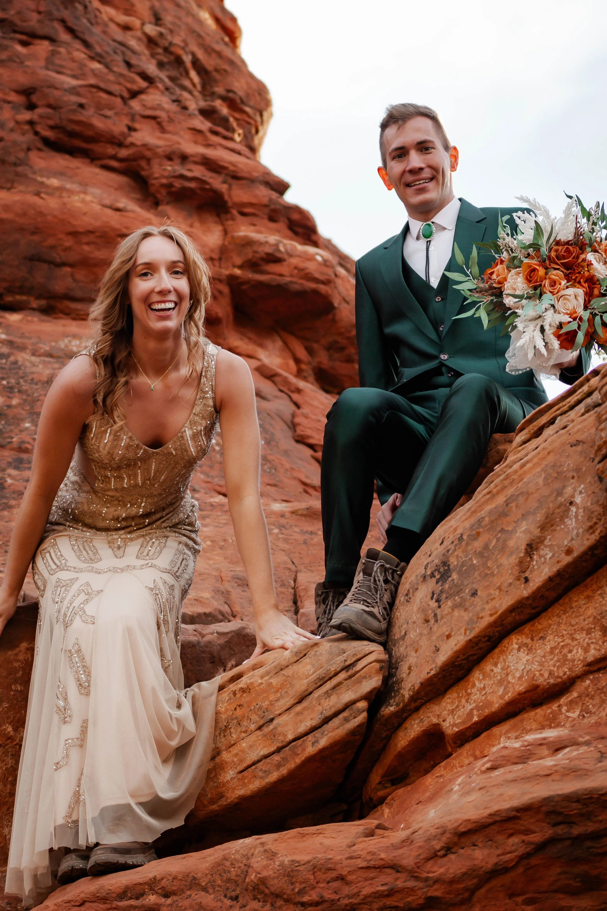 A smiling woman in a beige, beaded gown and a man in a green suit with a white shirt and bolo tie, holding a bouquet of flowers, pose on red rock formations outdoors.