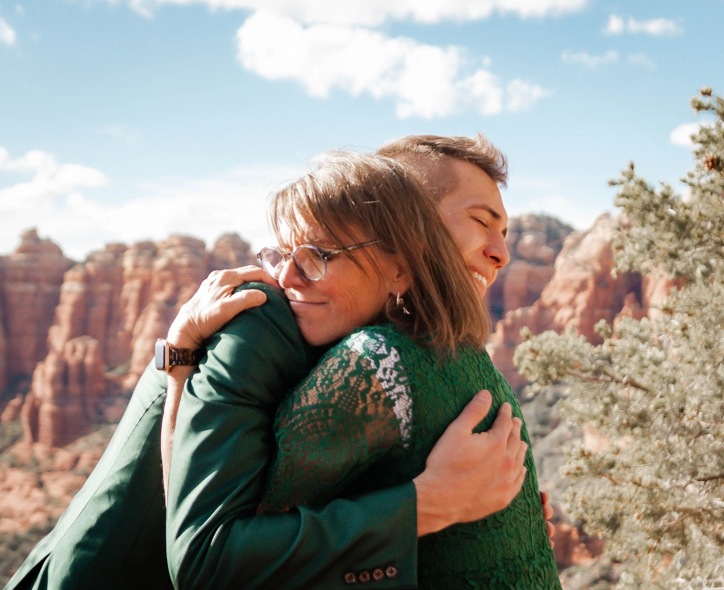 Two women hugging outdoors with a rocky, desert landscape and blue sky in the background.