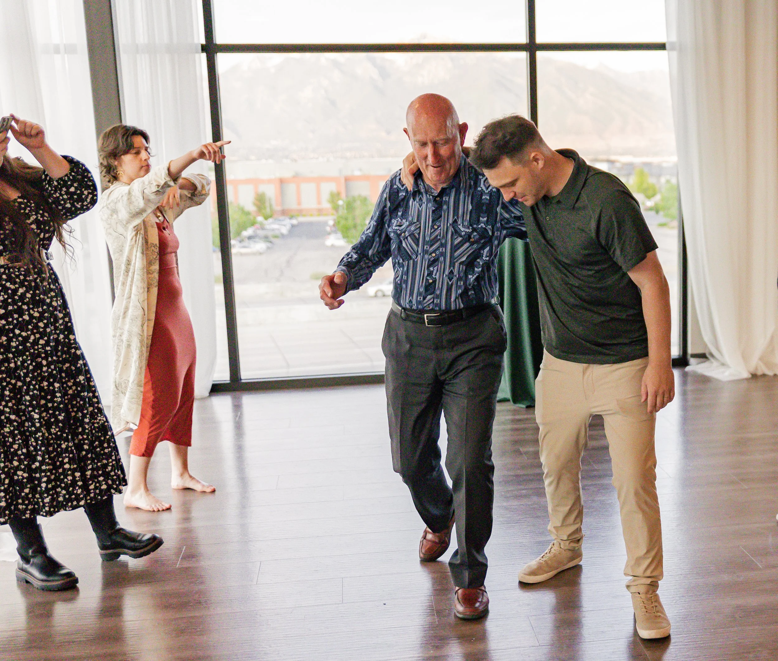 An elderly man and a young man dance together in a room with large windows, with other women in the background enjoying the moment.