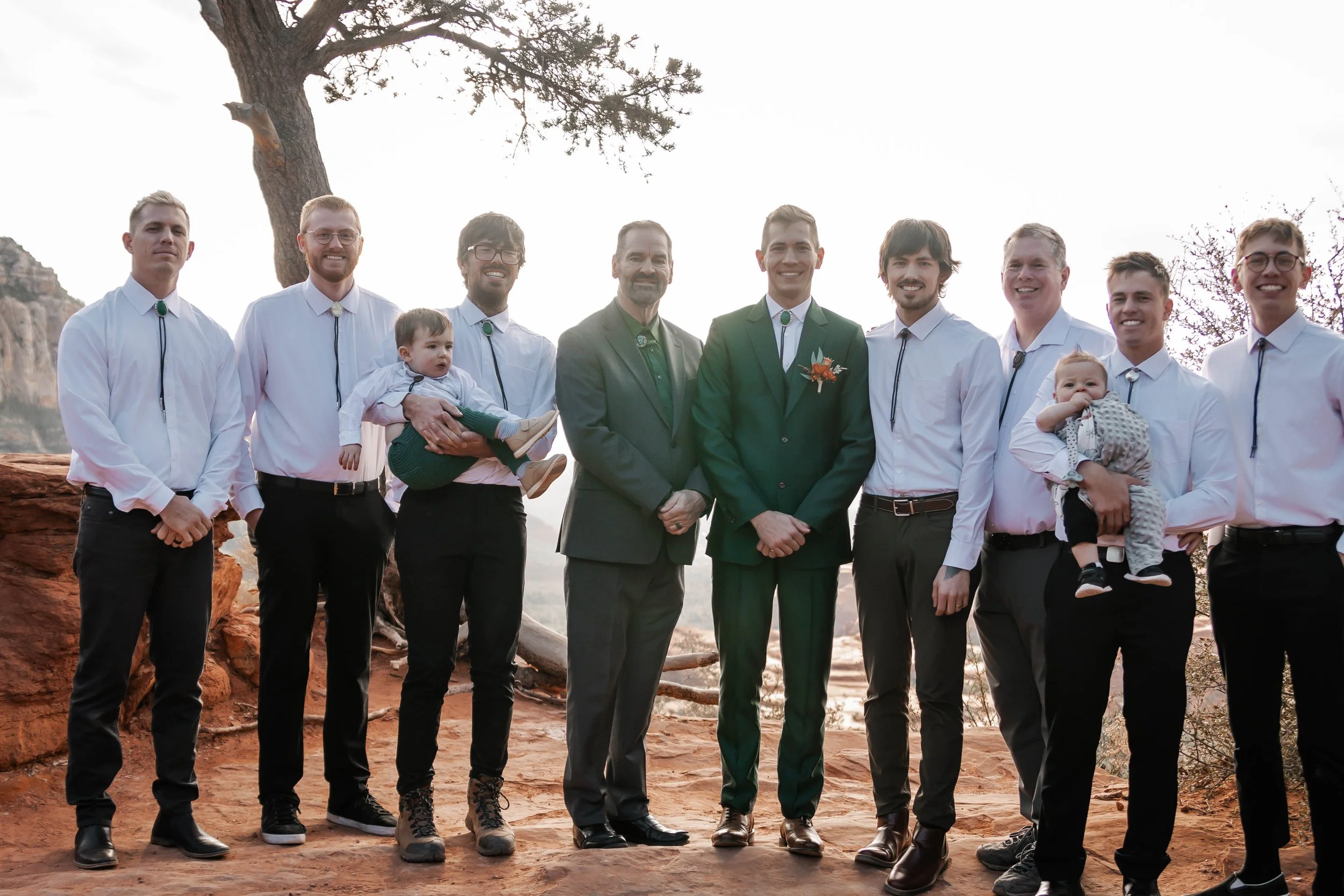 A group of people outdoors during sunset, including men in white shirts and two in suits, with two children, standing on red dirt with a tree and rock formations in the background.