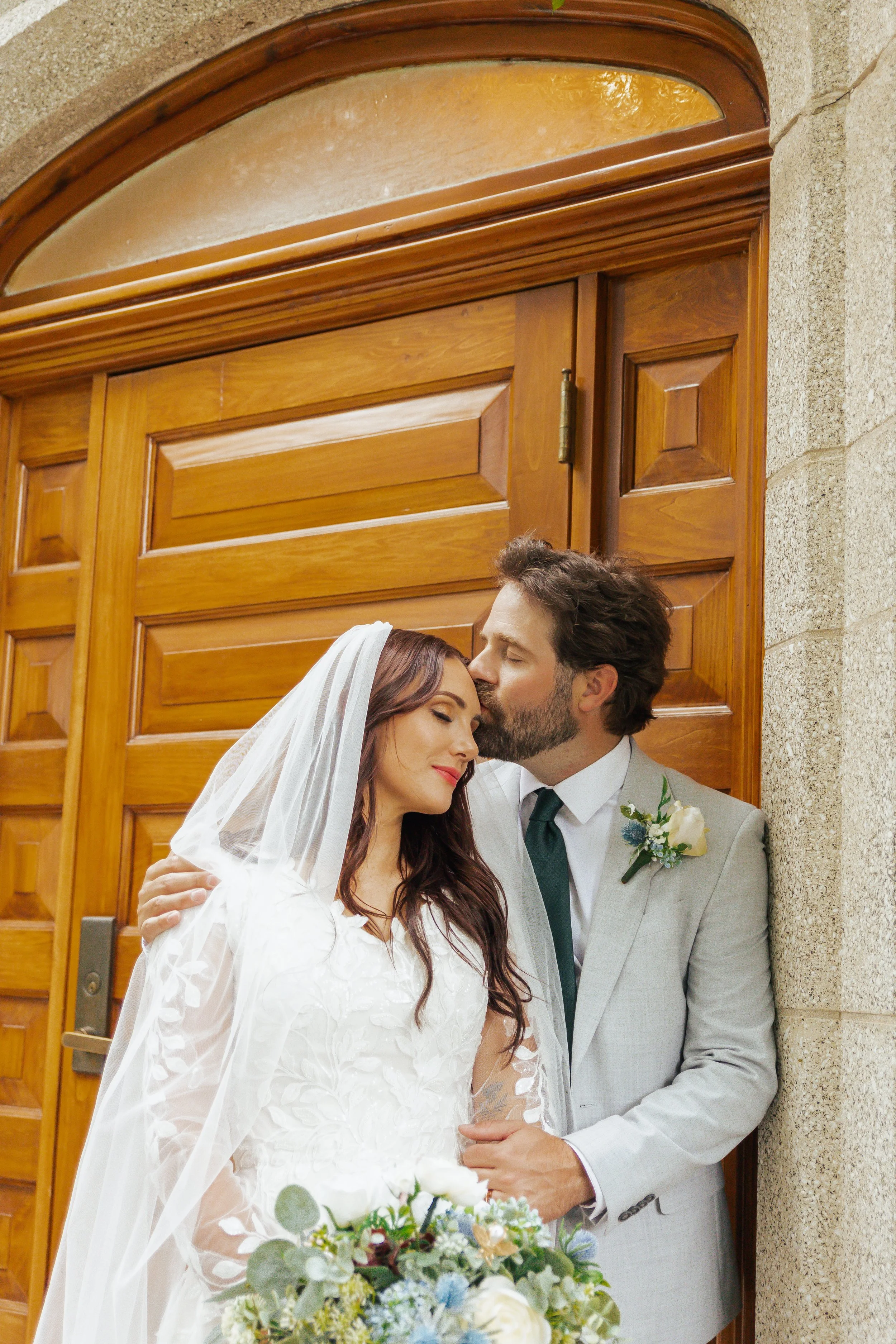 A bride and groom sharing a tender moment outside a wooden door, with the groom kissing the bride's forehead as she smiles softly, holding a bouquet of flowers.